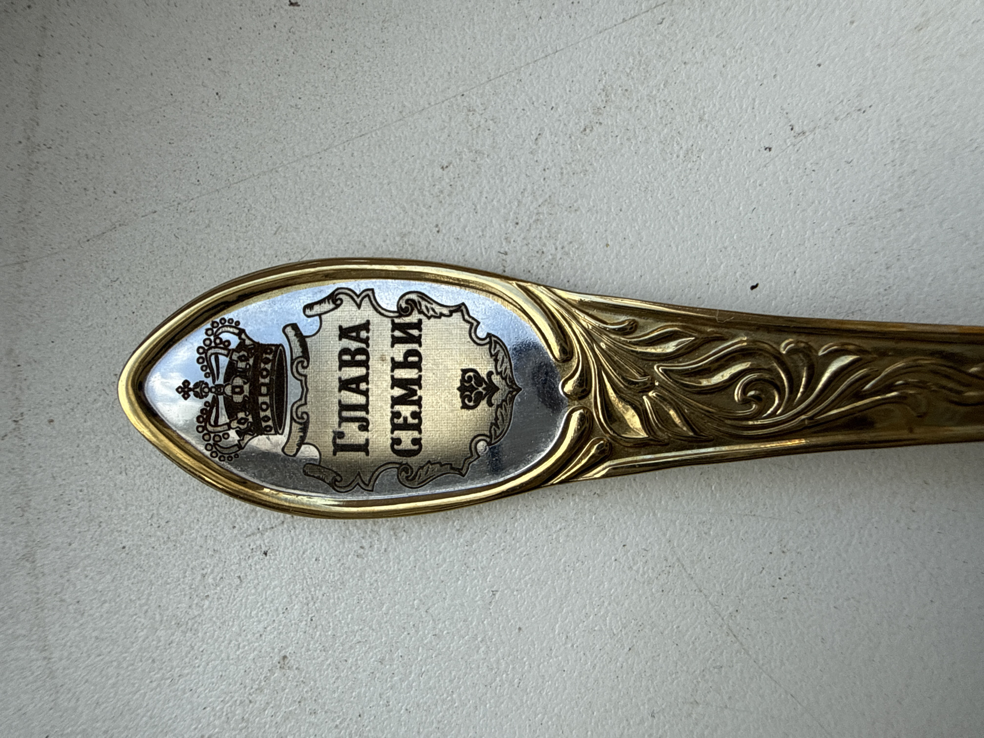 This media file shows a close-up, top-down view of the decorative end of a golden spoon. The spoon's handle features ornate, swirling patterns in gold. The bowl of the spoon is oval-shaped and edged with gold. Inside the bowl, a silver inlay contains black text and decorative elements.
The text, written in Cyrillic script, reads "ГЛАВА СЕМЬИ", which translates to "Head of the Family". To the left of the text is an illustration of a crown with intricate details, and to the right, a smaller, simpler crown-like symbol. The background of the spoon's bowl is a light, reflective surface. The spoon rests on a textured, light-colored surface, possibly a table or countertop. The lighting suggests it is daytime, with natural light casting subtle shadows. No people are visible in the image.