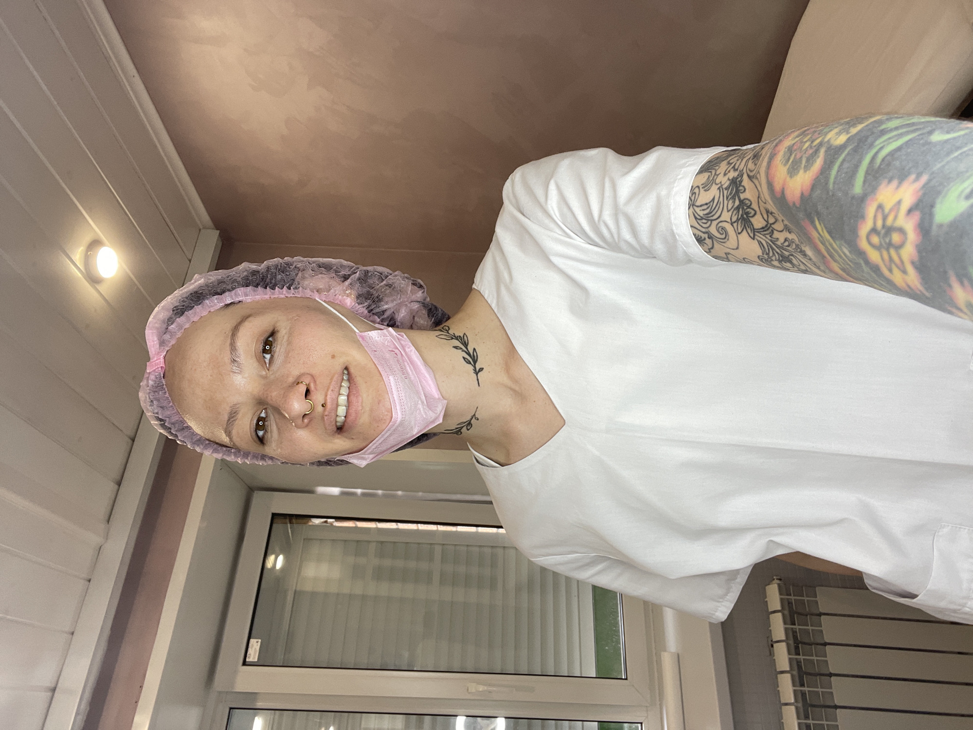 A person wearing a pink surgical cap and face mask, along with a white garment, is facing the camera. They have visible tattoos on their arm and neck. The background includes a window with blinds, a radiator, and a wall with a light fixture. The image might be taken inside a clinic. There is no visual information in this image to support the location is in the city of Izhevsk, Russia.
