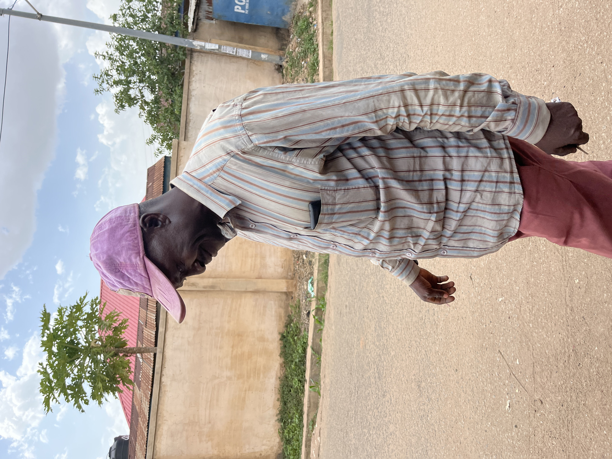 A man wearing a purple baseball cap, a long-sleeved striped shirt, and maroon pants is walking on a paved road. He appears to be in Jos, Nigeria, judging by the architecture, vegetation, and road quality, although precise location determination within the city is not possible with only this image. A building with a red roof and a tree in front of it is visible. Other buildings and trees are also present, suggesting a mixed urban and natural environment. The sky is partially cloudy.