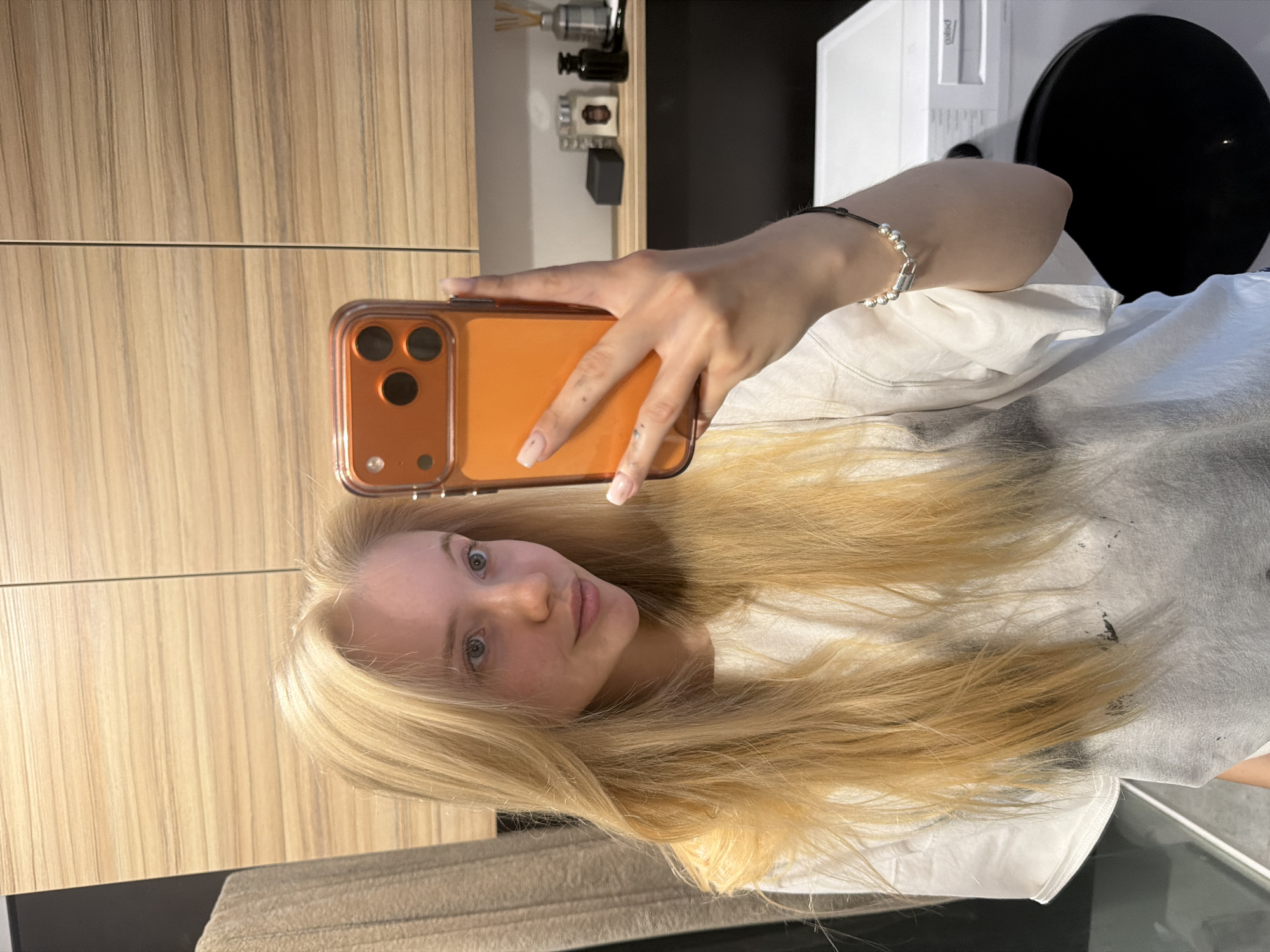 A person with long, blonde hair is taking a selfie with an orange-cased phone. The background includes a wooden cabinet, a shelf with various items, and a white appliance with a black circular component. The person is wearing a white t-shirt and a bracelet. The location is unknown.