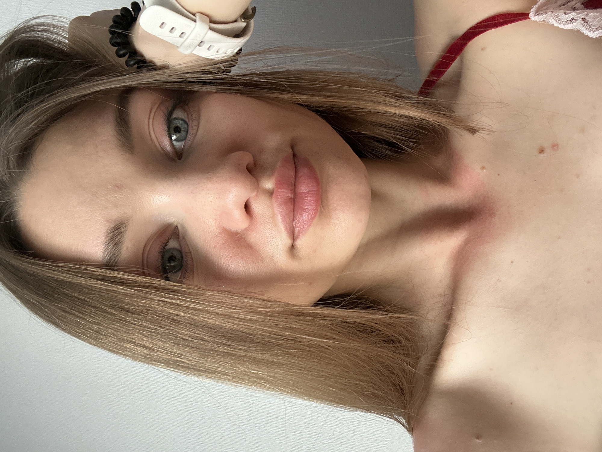 A close-up, selfie-style image of a light-skinned woman with light brown hair and greyish-blue eyes. She is wearing a white smartwatch on her wrist. The angle is slightly rotated, showing her face, part of her upper chest, and shoulder. She is wearing a red and lace strap. The background appears to be a plain wall, likely indoors. There is no information available that can pinpoint this picture was taken in the city of Kazan, Russia.