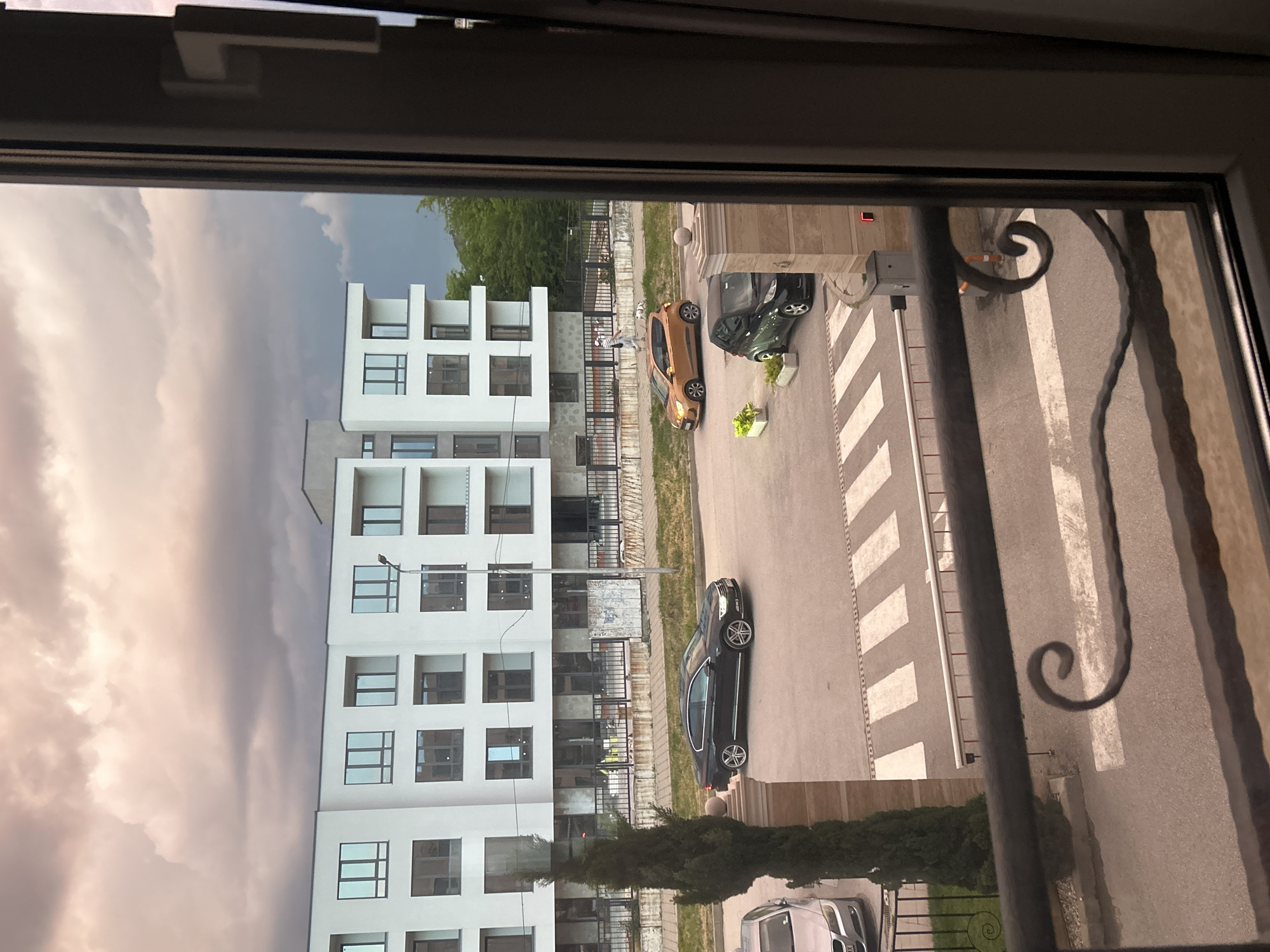View from a window, likely in an apartment, overlooking a street in Plovdiv, Bulgaria. The street is a crosswalk with white stripes. Several parked cars are visible. A modern, white building is to the left. Two people walking dogs are visible near one of the parked cars. The sky is partly cloudy. A decorative metal railing is visible on the right side of the window.