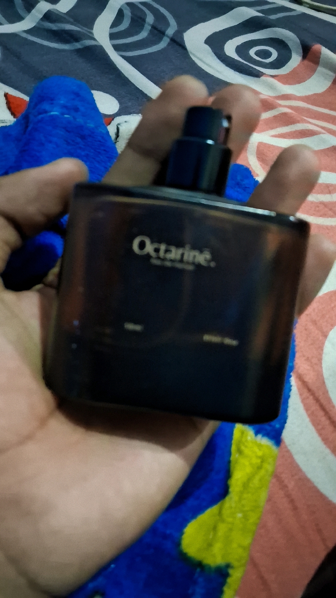 A person is holding a dark rectangular bottle of "Octarine" perfume. The bottle has a black cap and the brand name is in white text. The background is a blue blanket with yellow and pink patterned fabric. The scene appears to be indoors, possibly in a bedroom, given the bedding. There is no information in the image to suggest a specific location within Babakan, Indonesia.
