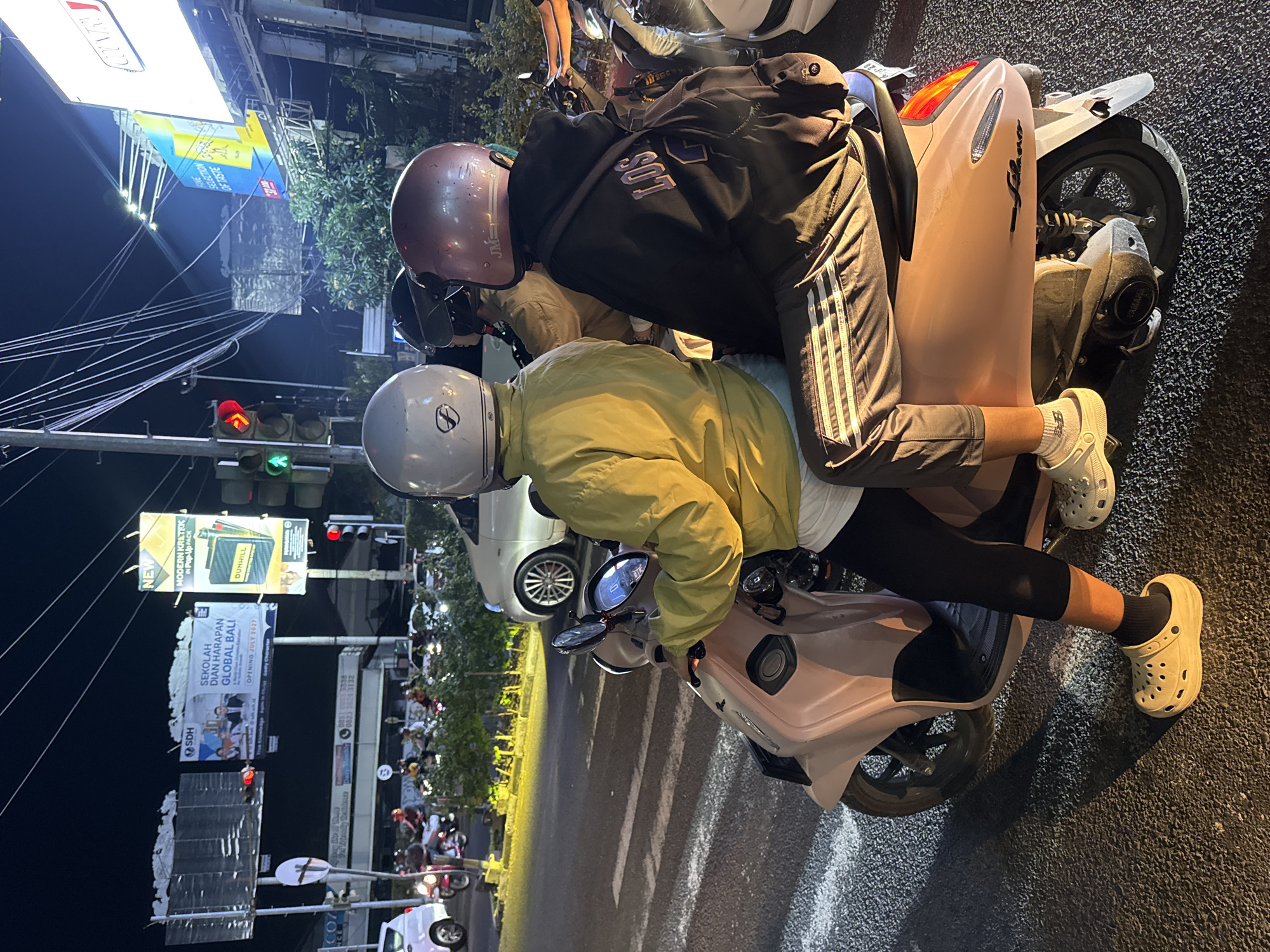 This media file captures a nighttime scene on a street in Denpasar Barat, Indonesia, likely during a traffic light stop. Two individuals are seated on a scooter. The person in front wears a yellow jacket and a silver helmet, while the person behind them is dressed in a dark hoodie and a maroon helmet. They appear to be riding together, with the person in front operating the scooter.
Several advertisements are visible in the background. One prominent billboard displays "MODERN KRETEK IN Pop-Up PACK" and "DUNHILL." Another banner reads "SEKOLAH DIAN HARAPAN GLOBAL BALI" with an opening date of "JULY 2017," suggesting an educational institution. There are also traffic lights, indicating an intersection. The street is asphalt, and the lighting suggests it is nighttime. The overall atmosphere is that of a typical urban street scene in Southeast Asia at night, with the presence of traffic and illuminated advertisements. Visible text includes brand names and information about a school.