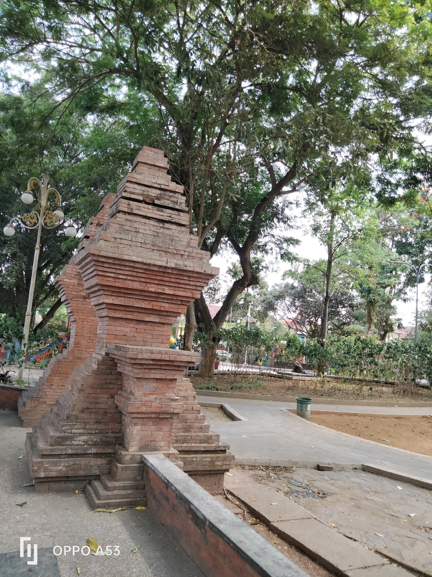 This is a vertical, eye-level shot of a brick structure at the entrance to a park in Trenggalek, Indonesia. The structure is made of reddish-brown bricks and is tiered, resembling a small temple or gateway. It is positioned on the left side of the frame, with its intricate brickwork prominently featured. Behind the structure, a lamppost with ornate golden accents and three white globes stands tall.
The background reveals a lush park environment. Large trees with dense green foliage dominate the upper portion of the image, with sunlight filtering through the leaves. A paved pathway winds through the park, leading towards the background. Various plants and bushes line the path, creating a natural and somewhat wild aesthetic. In the distance, parts of buildings with reddish-brown roofs are visible, suggesting a residential or commercial area bordering the park. The ground is a mix of dirt and paved areas, with some scattered leaves. The overall atmosphere is serene and natural.
The bottom left corner of the image displays the branding "OPPO A53". There are no visible people or animals in the frame. The lighting suggests it is daytime, with natural light illuminating the scene.