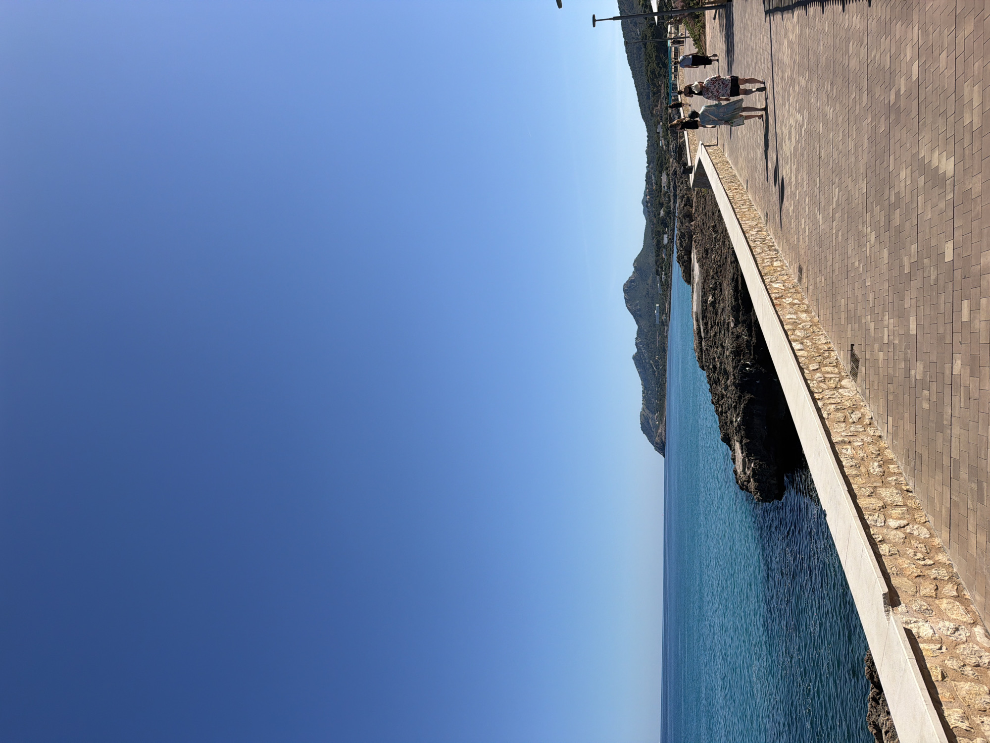 The image depicts a coastal scene in Capdepera, Spain. The sky is a clear, deep blue. The Mediterranean Sea is a lighter shade of blue. A rocky coastline with dark cliffs is visible. A paved promenade runs along the top of the cliffs, with a low stone wall separating the walkway from the drop to the sea. Several people are walking on the promenade. Tall light posts are spaced along the walkway. In the background, a sloping hillside is covered with trees.