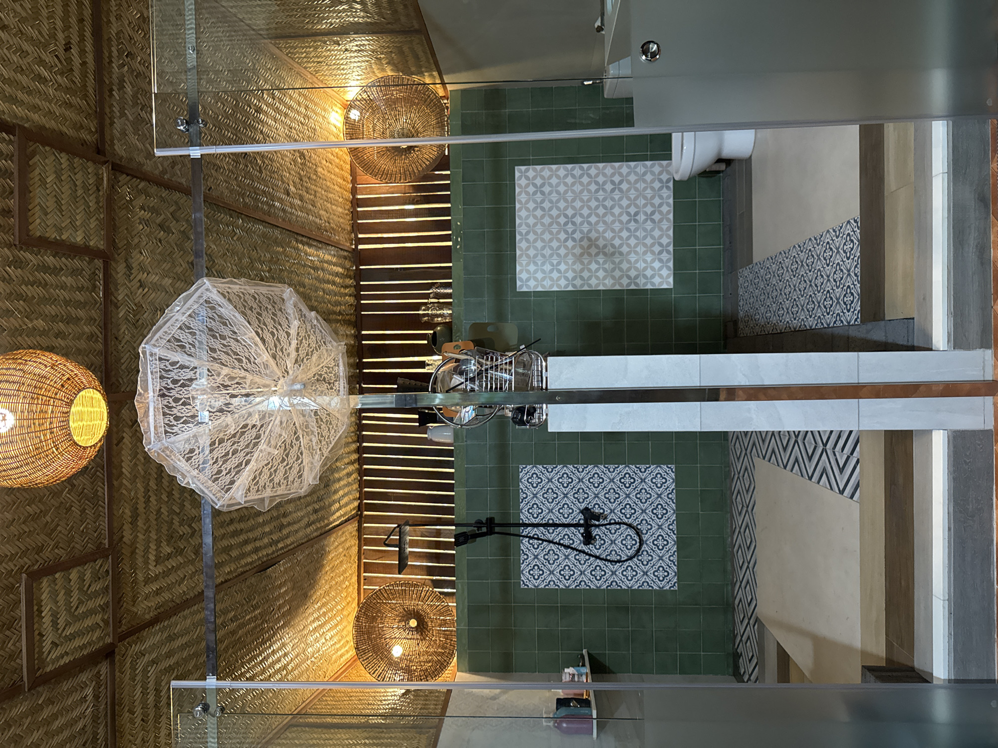This is a forensic, matter-of-fact description of a bathroom area within a resort in Mũi Né, Vietnam. The scene depicts two adjoining shower stalls separated by a reflective surface, with glass doors and walls. The ceiling is covered with woven natural materials, and two large, woven pendant lights hang from it, one visible in the upper center and another in the upper right.
The walls of the shower stalls are primarily tiled in a muted green, with decorative tile panels featuring geometric patterns on each side. The left stall has a black shower fixture and a small shelf holding several bottles. The right stall has a white toilet visible. The floor is a mix of tiled and wooden sections with decorative borders.
Two circular, woven wall sconces with visible light bulbs are mounted on the walls behind the shower areas, casting a warm glow. A decorative white lace umbrella is suspended from the ceiling between the two stalls. Vertical wooden planks line the back wall above the tile.
The overall impression is one of a tropical, potentially luxurious, bathroom design with natural materials and a focus on open shower spaces. There are no people visible in the image. The environment appears clean and well-maintained.