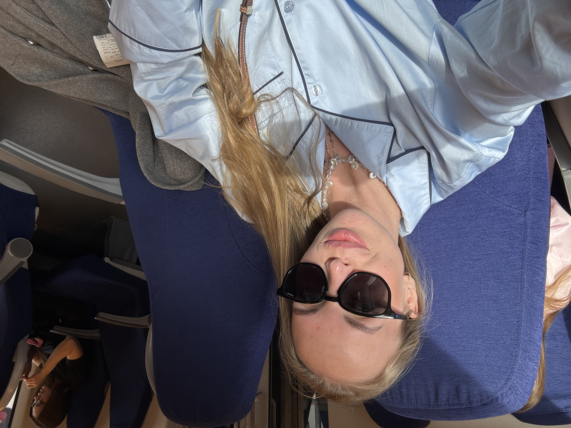 A woman is sitting in what appears to be an airplane or train seat. She is wearing sunglasses and a light blue, button-down, pajama-style shirt. Her hair is blonde and partially covers her shirt. She also wears a necklace. There is a gray coat or blanket next to her. The seat is upholstered in dark blue fabric. Another person can be seen in the background, also sitting in a seat. The setting suggests an enclosed space, possibly during travel. There is no explicit indication of Sochi, Russia.