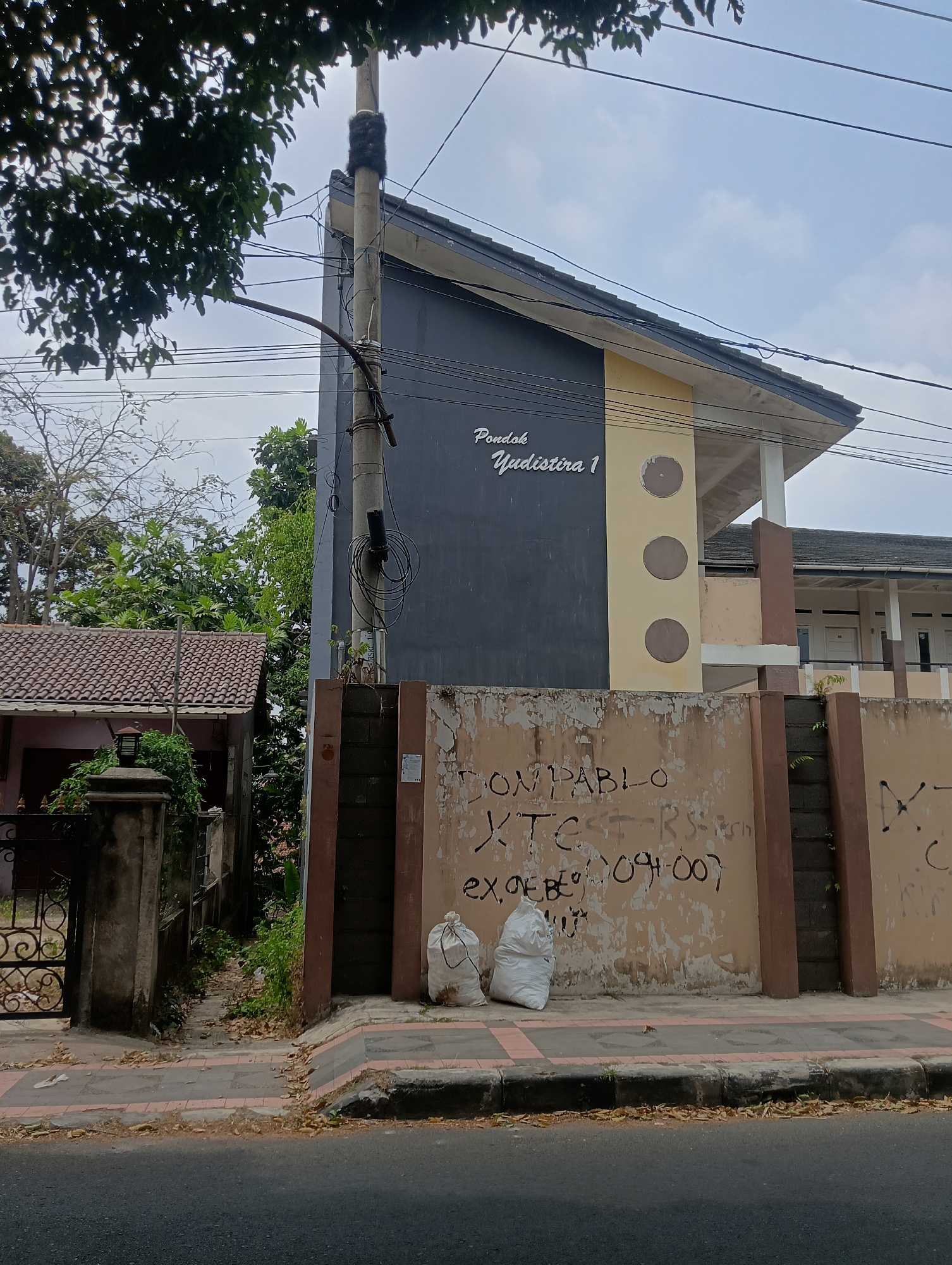 The image shows a street scene in Ciporang, Indonesia, featuring a building with a distinctive angular roof and a dark gray facade, accented with a yellow section with circular decorative elements. The building has "Pondok Yudistira 1" written in cursive script on the dark gray portion. In front of the building is a weathered, light brown wall covered in graffiti, including the phrases "DON PABLO XTC RS" and "EX, OEBO 094007", along with other illegible markings. Two white trash bags are placed on the sidewalk in front of the graffitied wall. To the left of the building, a house with a brown tiled roof and a dark metal gate is visible. The area is lined with trees and vegetation, and overhead power lines are present. The street in the foreground is paved, with a sidewalk separating it from the buildings. The sky is overcast.