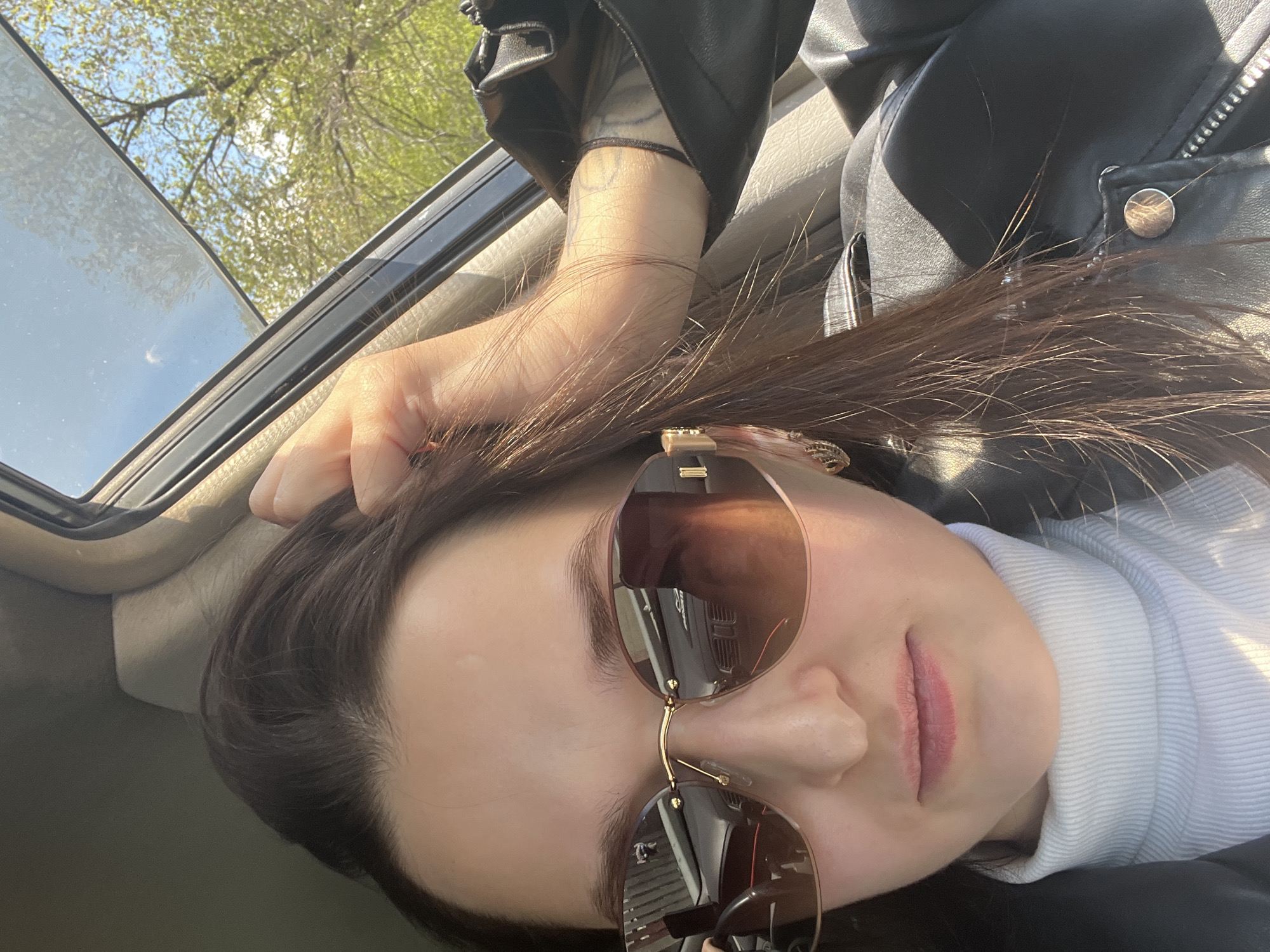 A woman is visible, wearing sunglasses, a white turtleneck shirt, and a black leather jacket. She is positioned in a car, evidenced by the sunroof. Outside the sunroof, trees are visible, suggesting an outdoor location. Based on the information provided, the location is the city of Dzerzhinsk, Russia, though no specific landmarks are discernible in the image. The woman's arm is raised, with her hand near her hair. The overall scene suggests the woman is relaxed and enjoying the sunlight in her car.