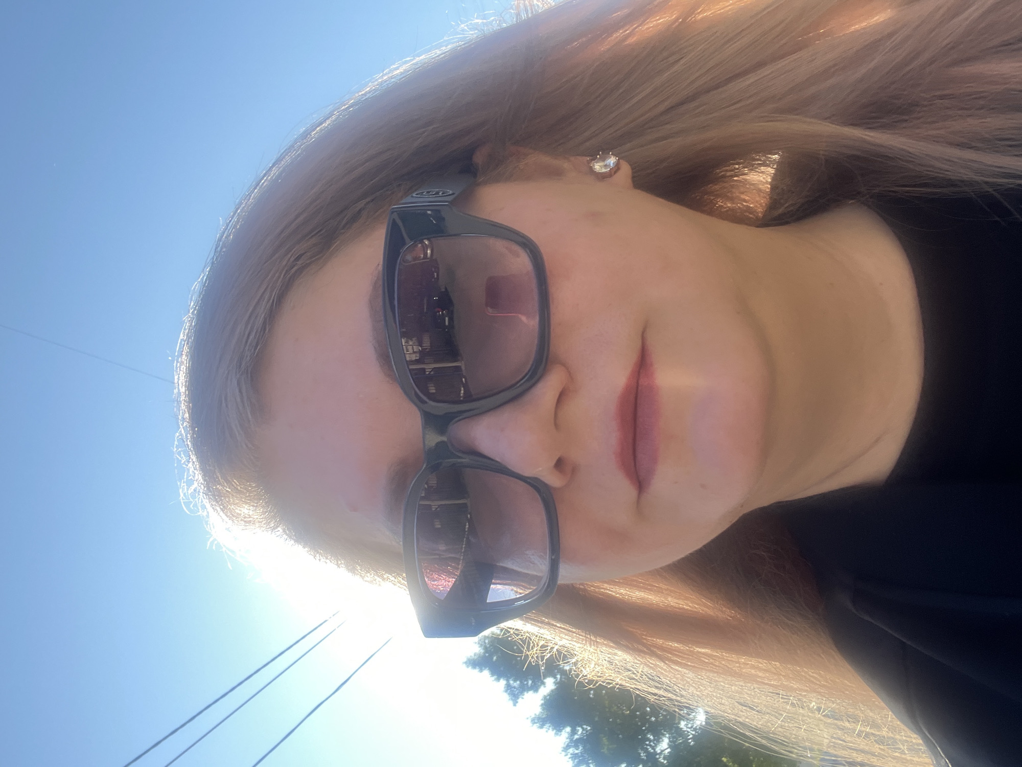 A woman with blonde hair, wearing black sunglasses and a black top, is captured in a close-up, outdoor, selfie-style photograph. Her face is illuminated by the sun, which is positioned behind her and to the left, creating a lens flare. She has on light pink lipstick and a diamond stud earring on her right ear. The background consists of a clear blue sky with power lines visible in the upper left quadrant, and trees to the left. The overall setting appears to be a sunny day in a suburban or park-like area. The image does not provide any specific clues to pinpoint its location within Izhevsk, Russia.