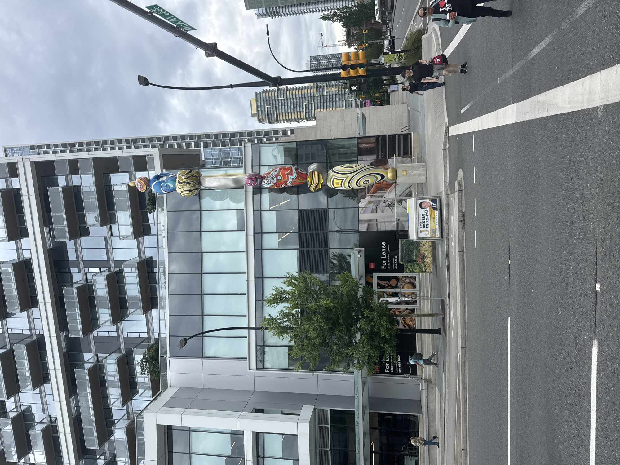 An outdoor, eye-level, full shot captures a busy street scene in Burnaby, Canada. A large, modern building dominates the left side of the frame, its glass facade reflecting the cloudy sky. A colorful totem pole-like sculpture stands tall in front of the building. In the foreground, a street with white lane markings runs across the bottom of the image. Pedestrians are visible on the sidewalk and crossing the street. Traffic lights and street signs are present, indicating a public intersection. The overall atmosphere is that of an urban environment on a slightly overcast day.