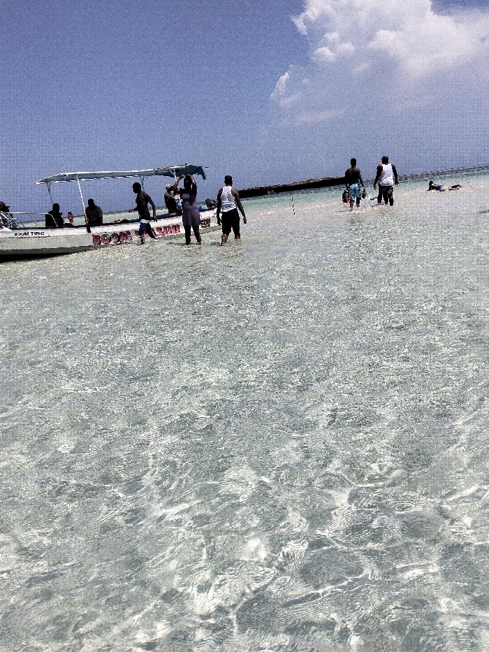 The image depicts a bright, clear coastal scene in Wasini, Kenya, dominated by shallow, rippled water extending from the foreground. A white boat with a light-colored canopy is positioned in the midground, left of center. Red text on the boat's side identifies it as "BOOM WASINI" and "MAJIN TURO". Approximately seven individuals are visible wading in the transparent water. Four individuals are in close proximity to the boat: one person walks left near the bow, another stands facing right with arms raised towards their head, and two others are standing or walking in the water near the stern. At least three additional individuals are seated inside the boat under the canopy. Further to the right in the midground, two more individuals are present in the water; one is standing, and another is lying prone. The background consists of a low, dark horizontal landmass or barrier under a clear blue sky with scattered white cumulus clouds.