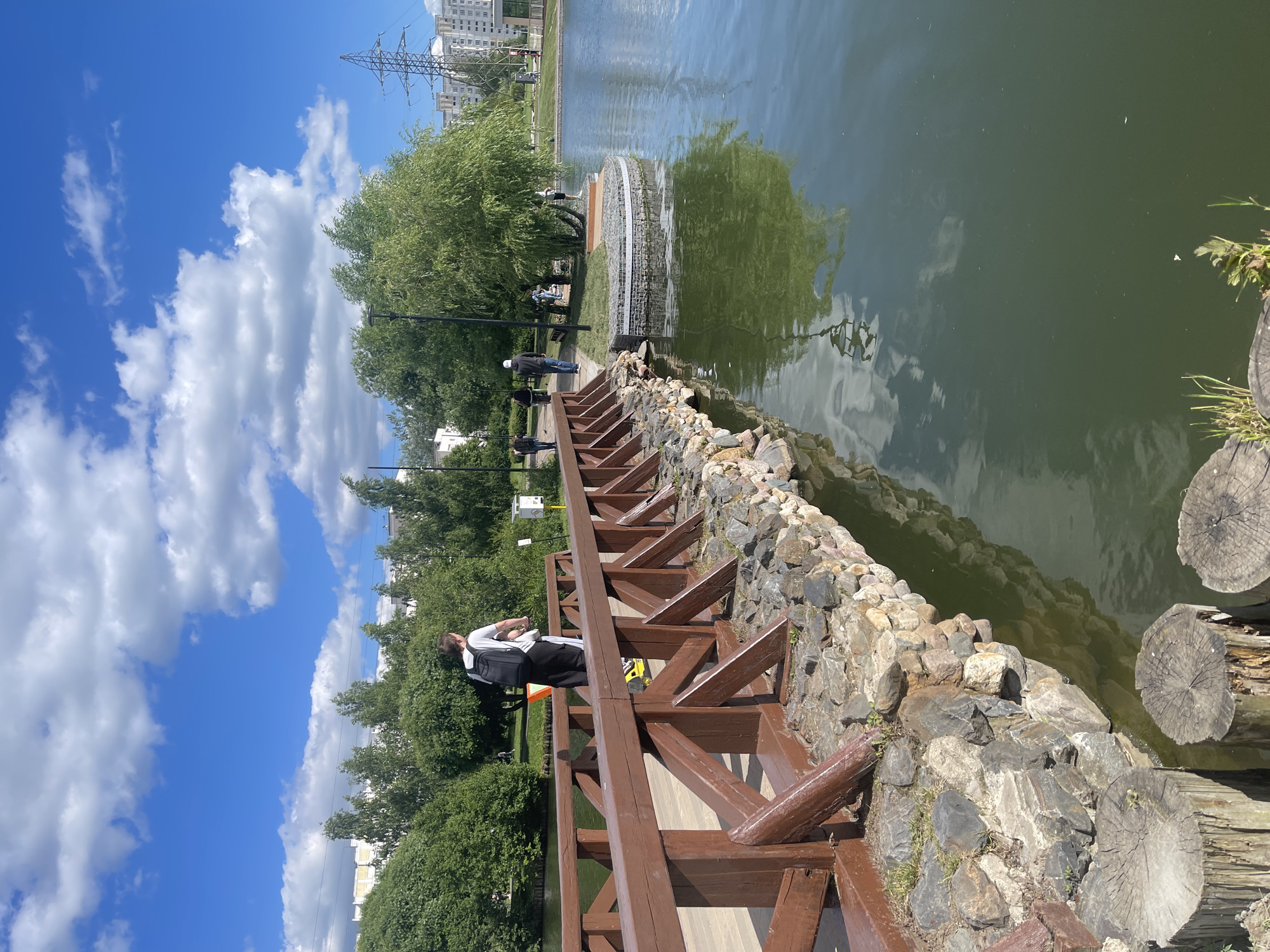 The image depicts a park scene with a wooden bridge over a body of water. Several individuals are visible walking on the bridge and on the grassy area near the water's edge. The environment includes lush green trees, a cloudy blue sky, and a cityscape in the background, featuring apartment buildings and a power line tower. The bridge is constructed with dark brown wood and has a stone embankment. The water appears calm with reflections of the sky and surrounding trees. The overall impression is that of a recreational area in an urban setting.