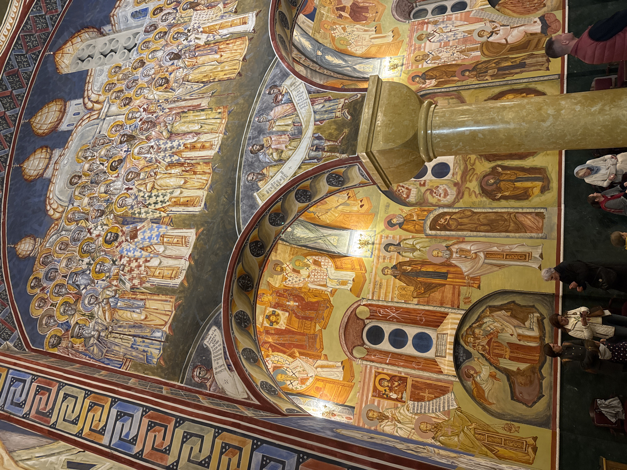 The image shows a portion of the interior of the church of St. John Vladimir in Bar, Montenegro, specifically the ceiling and upper walls. The surfaces are covered in detailed, colorful Orthodox Christian frescoes. The images depict various religious figures, scenes from scripture, and groupings of saints, distinguished by golden halos. A substantial, cylindrical column bisects the composition, obscuring some of the mural. Several people can be seen looking up at the frescoes in the periphery of the image.