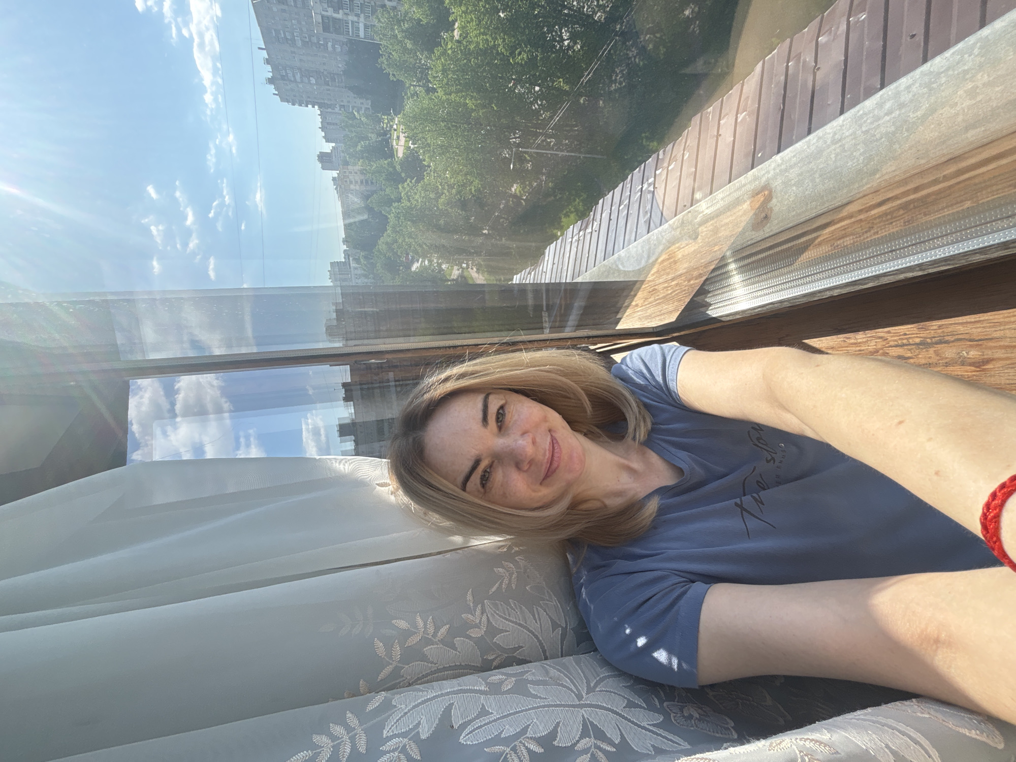 A woman is lying down on a sofa or daybed positioned next to a window. She is wearing a short-sleeved blue t-shirt. The woman is fair-skinned and appears to be blonde. She is smiling slightly at the camera. Outside the window, the image reflects the buildings and trees, suggesting an urban environment with a sky visible. The buildings indicate it is likely a city or urban location. The exterior structure of the window frame appears to be made of wood. The curtain is light-colored and possibly lace or embroidered, with a leaf pattern. She appears to be wearing a red bracelet.
