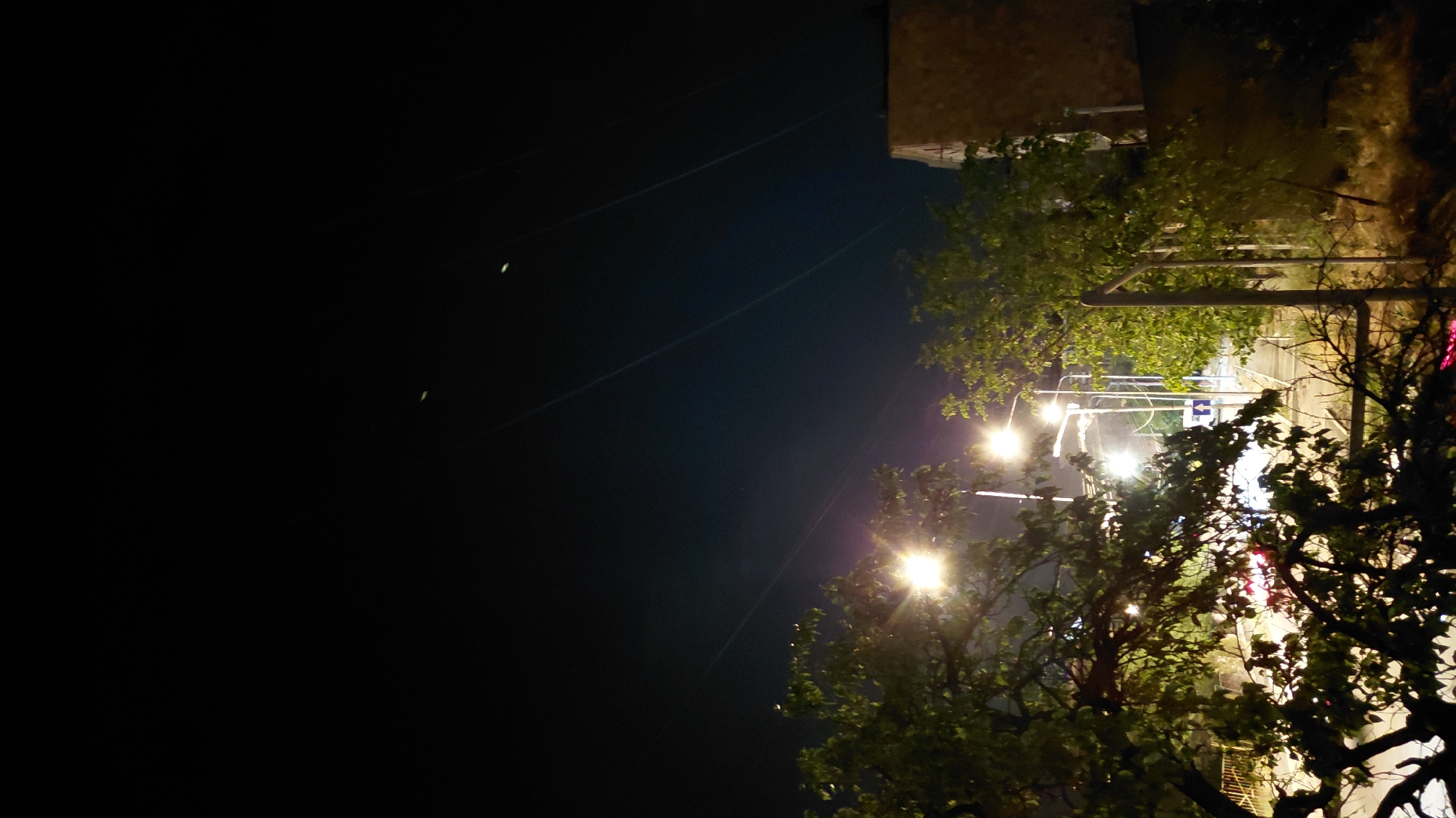The image depicts a nighttime scene with streetlights illuminating trees and a building in Abovyan, Armenia. The sky is dark, with several faint, linear light trails, possibly from aircraft or distant lights. In the foreground, leafy branches of trees obscure parts of the view. Streetlights with bright yellow illumination are visible, casting light on the surrounding foliage and a street below. The street appears to have traffic, with some red light streaks indicating vehicle movement. A building with a brick facade is partially visible on the right side of the image. The overall atmosphere is nocturnal, with artificial lighting from the street and the building providing the primary illumination.