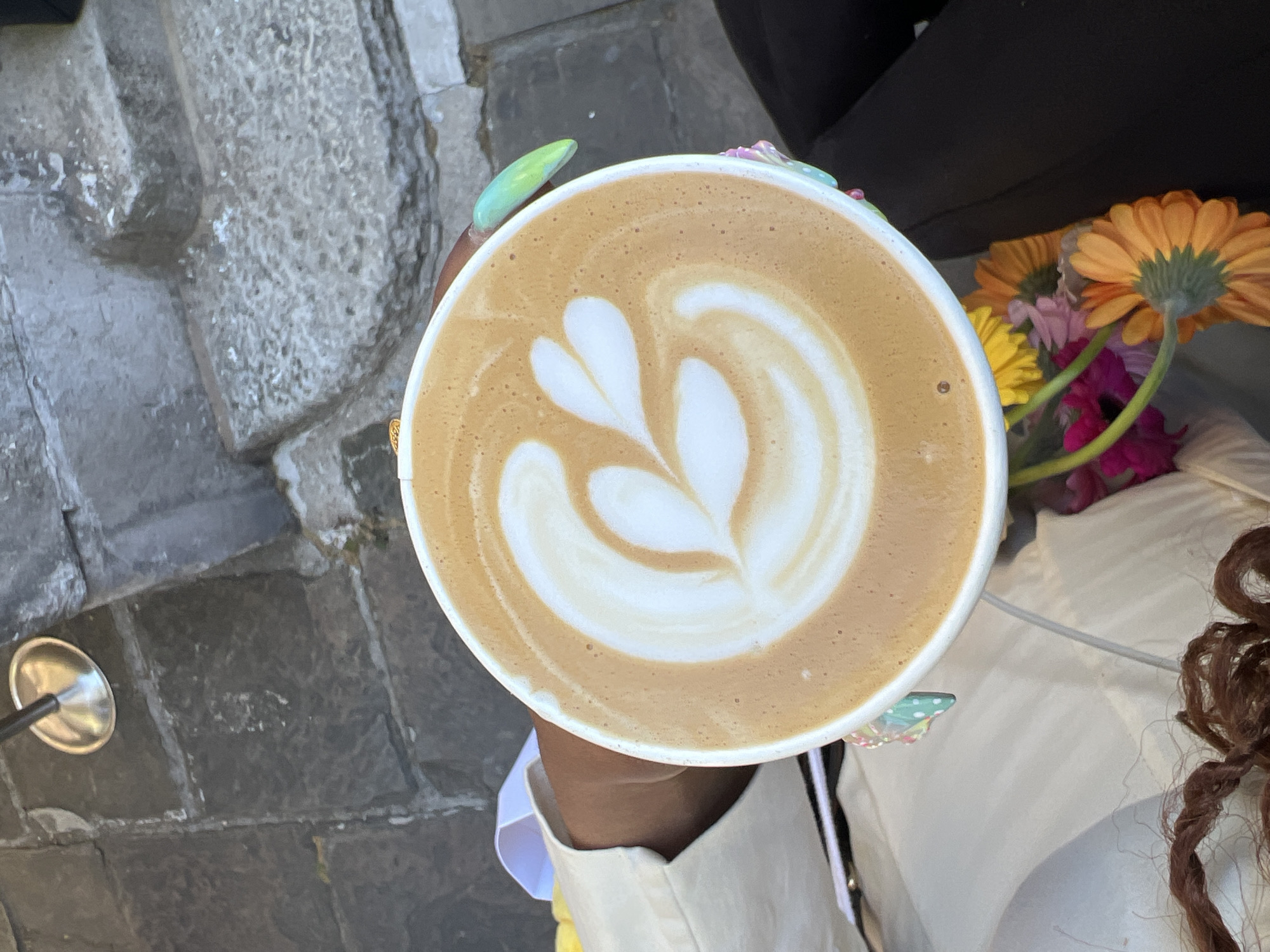 An overhead shot captures a person holding a white disposable coffee cup outdoors in what appears to be London, United Kingdom. The cup is filled with a light brown beverage, likely coffee or a latte, featuring intricate white latte art in a classic heart/leaf pattern.
The person's hand, arm, and part of their torso are visible, dressed in a light-colored top or jacket. Their fingernails are distinctively styled; one visible nail on the left is a vibrant lime green, while others feature colorful butterfly or whimsical designs. To the right of the coffee cup, a small bouquet of fresh flowers is visible, including prominent orange gerbera daisies, along with yellow, pink, and magenta blooms, suggesting a pleasant, possibly cafe-like setting. Parts of the person's brown, curly or braided hair are also visible towards the bottom right.
The background consists of textured grey paving stones or cobblestones, typical of European city streets or patios, hinting at an outdoor venue. On the far left, a small, reflective metal object, possibly the base of a stand or furniture leg, is seen on the ground. The bright, even lighting suggests it is daytime and likely sunny, contributing to a relaxed and enjoyable atmosphere. No discernible text is visible in the image.