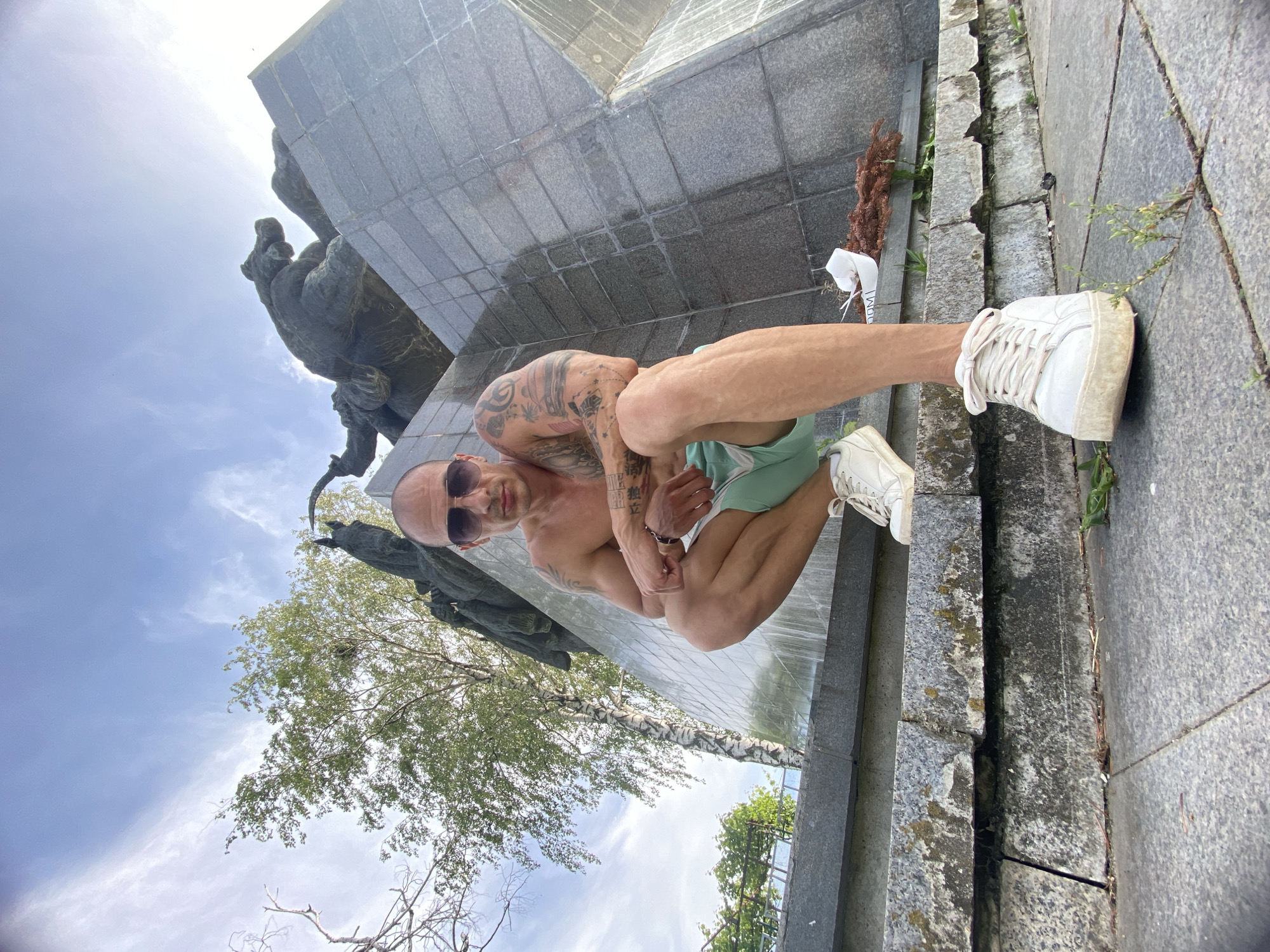 A man is squatting on a stone platform in front of a monument. He is shirtless, wearing sunglasses and light-colored shorts. He has tattoos on his arms. The monument appears to be made of granite and features bronze sculptures. A tree with green leaves is visible behind the man and to his left. The sky is blue with some clouds. The ground is paved with stone tiles. This is the Monument to the Soviet Army in Pleven, Bulgaria, also known as the Monument of the Liberators.