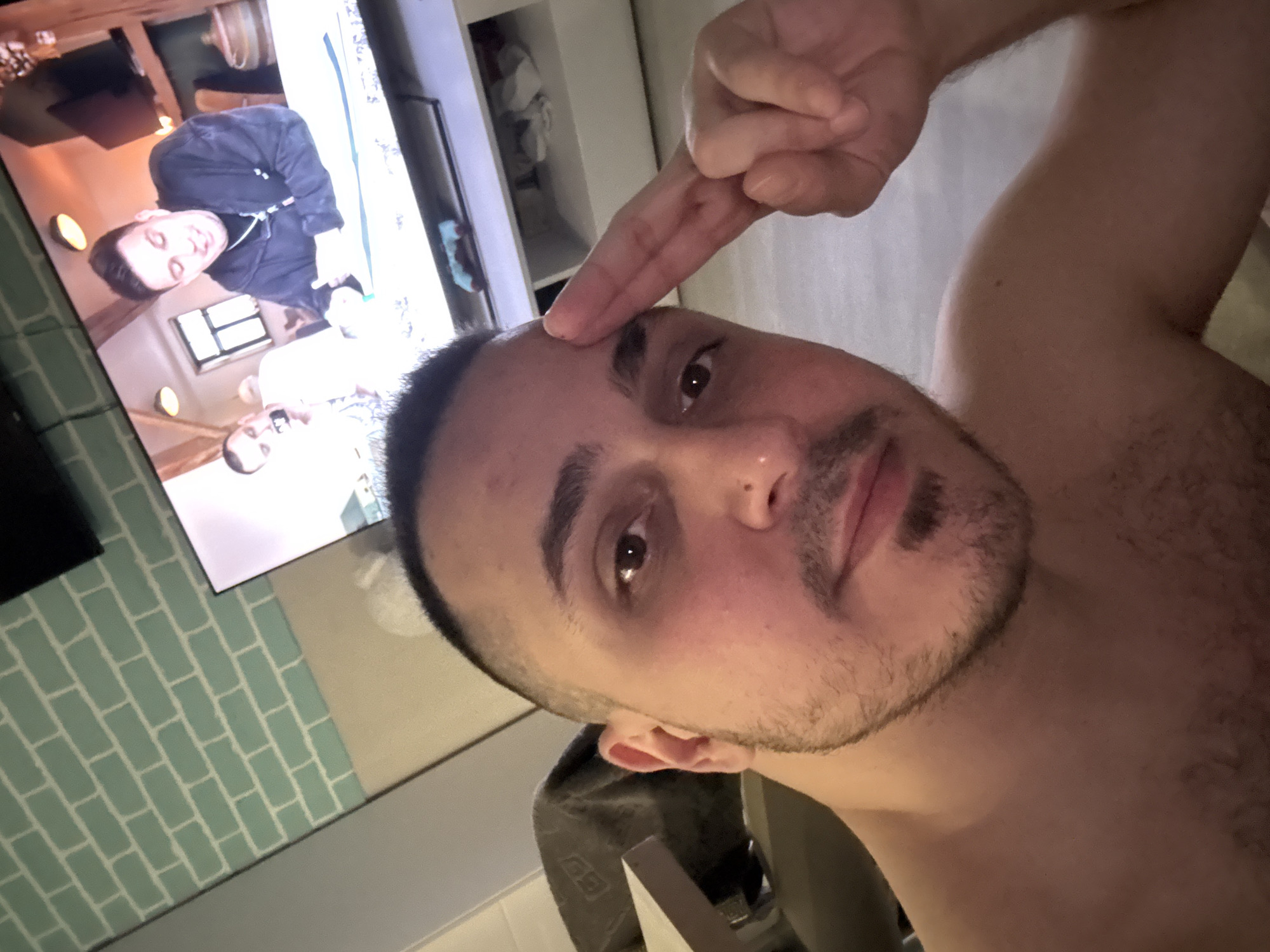 The image displays a man taking a selfie indoors, oriented vertically with the man's head on the left and his body extending to the right. The man, who appears to be in his 20s or 30s, has short dark hair, stubble, and dark eyes. He is shirtless and looks directly at the camera with a neutral expression. His right hand is raised, with fingers pressed together, touching his forehead in a gesture that resembles a salute or a point.
In the background, a large flat-screen television is visible, displaying content featuring at least two men. One man on the TV, wearing a dark hooded top, is clearly visible, looking to his left with a slight smile. Below him, another man is partially visible. The wall behind the television is adorned with a light greenish-blue subway tile pattern. Other indistinct furniture or shelving units are present in the background. The indoor lighting suggests the photograph was taken during the evening or night. There is no visible text in the image.