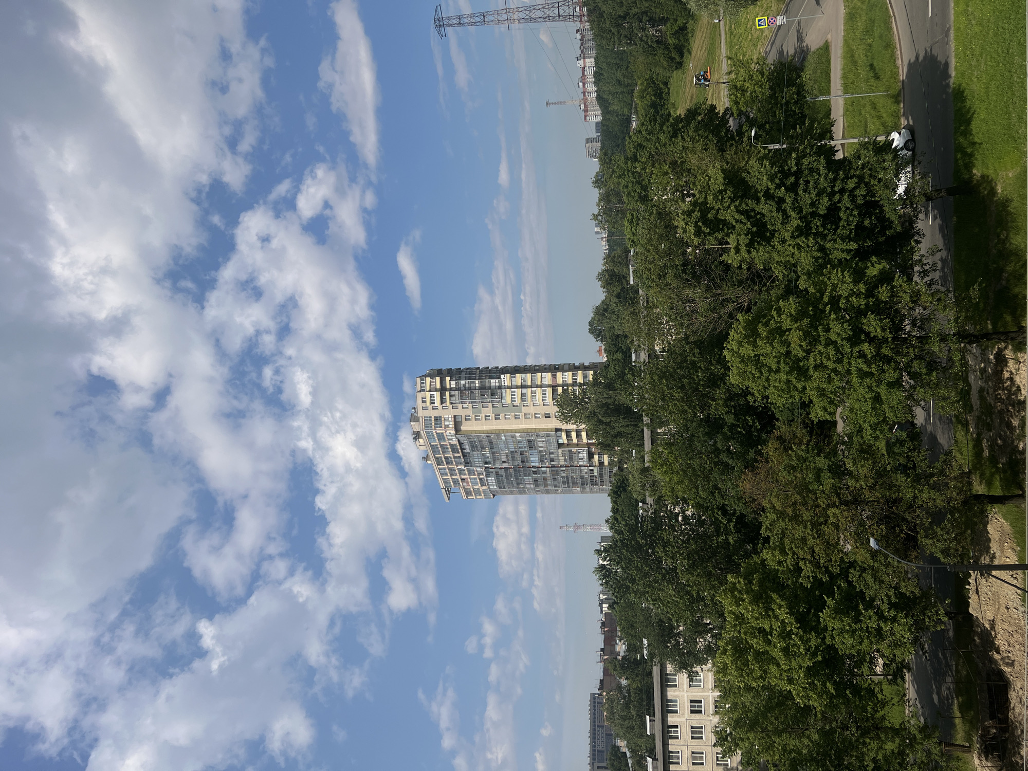 The image depicts an urban landscape under a bright, cloudy sky. In the foreground, dense green trees dominate, with a road curving through the scene. A white car is visible on the road. A street lamp stands tall among the trees. Further back, several apartment buildings are scattered amongst the foliage, with one notably tall, multi-story building with a distinctive modern design featuring many windows. Beyond the buildings and trees, a high-voltage power line tower stands in the distance. The location is identified as Petro-Slavyanka, Russia.