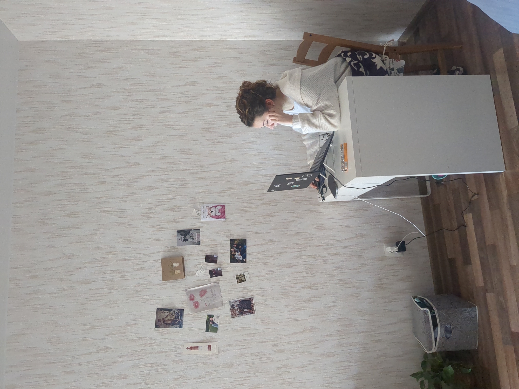 A person is seated at a desk with a laptop in a room with wallpaper. The desk is white, and there are various objects on and around it, including pens, a notebook, and wires. The person is wearing a white cardigan and is looking at the laptop screen. There are several small pictures and cards attached to the wall next to the desk. The room appears to be a workspace or home office. There is a wooden chair behind the desk, and a patterned storage box and plant are located near the floor. The floor is wood laminate. The location is unknown and it is unknown whether it is in Saint Petersburg, Russia.