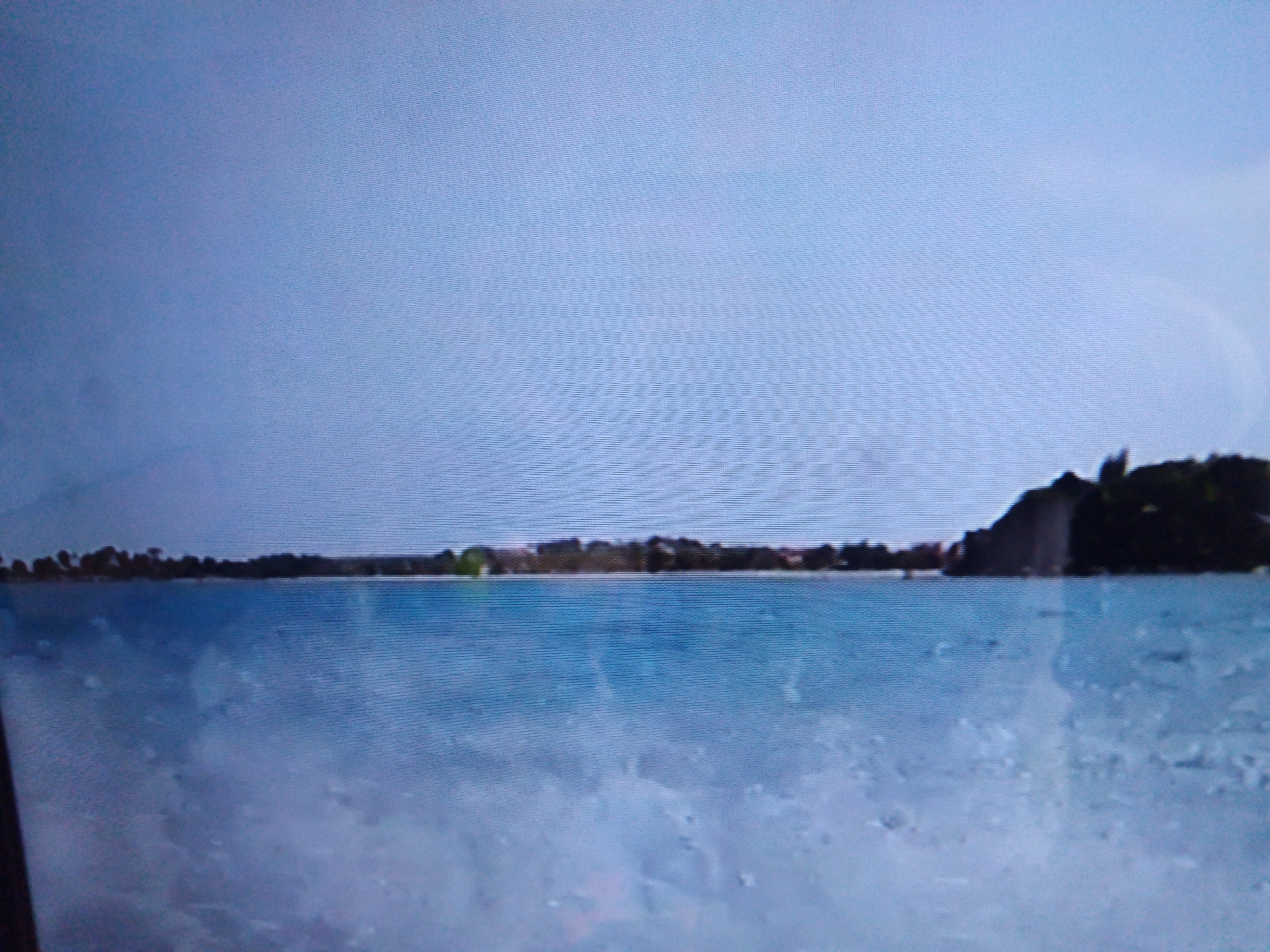 The image depicts a tranquil scene of a body of water under a pale blue sky. The water surface is calm, reflecting the soft light. Along the horizon, a distant shoreline is visible, lined with trees and some structures that appear to be buildings. To the right, a dark, hilly landmass rises from the water, covered in vegetation. The overall impression is one of natural beauty and serenity. Based on the information provided that this image is located in the city of Dushanbe, Tajikistan, this scene is likely depicting a reservoir, lake, or a large section of the Dushanbe sea, possibly one of the many water bodies and recreational areas within or surrounding the capital city.