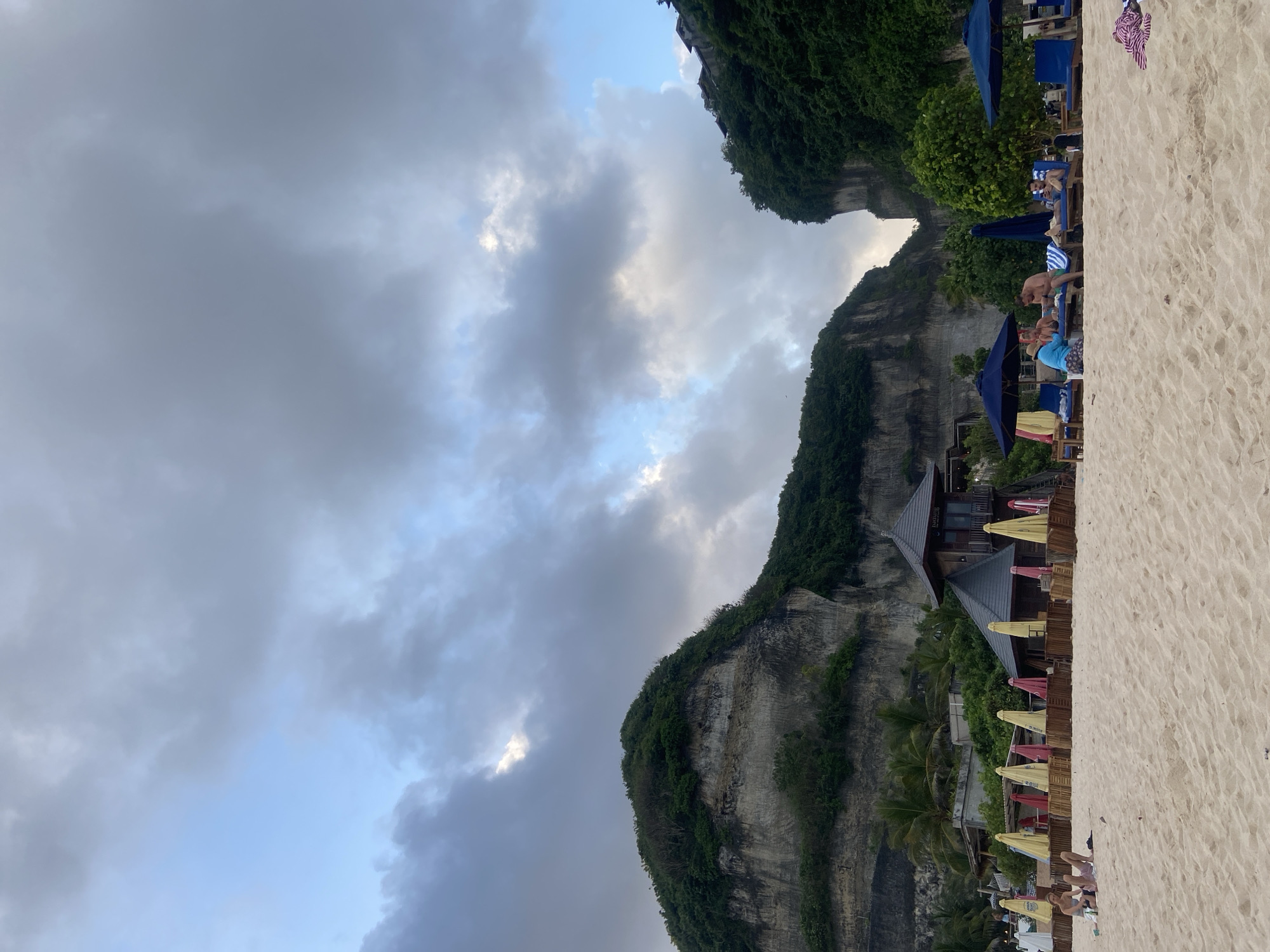 The image depicts a beach scene in Kuta Selatan, Indonesia, where a sandy beach meets a steep, rocky cliff face covered in vegetation. The sky is overcast with dark clouds. A structure with a wooden facade and multiple roofs is nestled at the base of the cliff. Beach umbrellas in varying colors (yellow, red, and blue) and lounge chairs are arranged along the beach. People are seen relaxing on the beach and on the lounge chairs under the umbrellas. The beach sand takes up a portion of the frame.