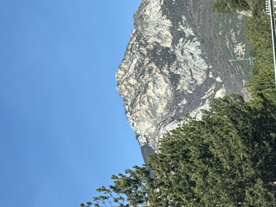 The image displays a majestic natural landscape featuring a prominent mountain face, a clear blue sky, and dense green foliage. The original image appears to be rotated 90 degrees clockwise; when viewed in its intended vertical orientation (rotated counter-clockwise), the scene becomes clearer.
In this vertical orientation:
On the right side of the frame, a large, imposing rock face of a mountain dominates. This rock face is a mix of light grey, white, and beige tones, with intricate textures, shadows, and crevices suggesting its ancient, weathered form. A notable feature of this mountain is the striking resemblance of its left profile to a human face. This natural formation clearly defines a forehead, a prominent nose, and a distinct chin, all looking towards the left into the clear sky.
The left side of the image is filled with a vast expanse of bright, clear blue sky, devoid of clouds, indicating fair and sunny weather.
In the foreground, particularly across the bottom of the image and extending up the lower left side of the mountain, is a dense mass of dark green trees. These appear to be coniferous, with their thick foliage creating a rich, textured foreground against the rugged mountain and bright sky. Further up the mountain, beyond the main rock face, slopes are covered in lighter green vegetation.
Minimal man-made elements are present:
Towards the top right portion of the mountain, against a backdrop of green foliage, a thin white pole with a small, dark, rectangular object (possibly a solar panel or sensor) attached is visible. In the very top right corner, a segment of a white vertical structure, possibly a pole or part of a distant building, can be seen.
The time of day is clearly daylight, likely mid-day, given the bright illumination and clear sky. There is no visible text in the image. The most distinctive aspect of the image is the natural pareidolia, where the mountain's profile strikingly resembles a human face.