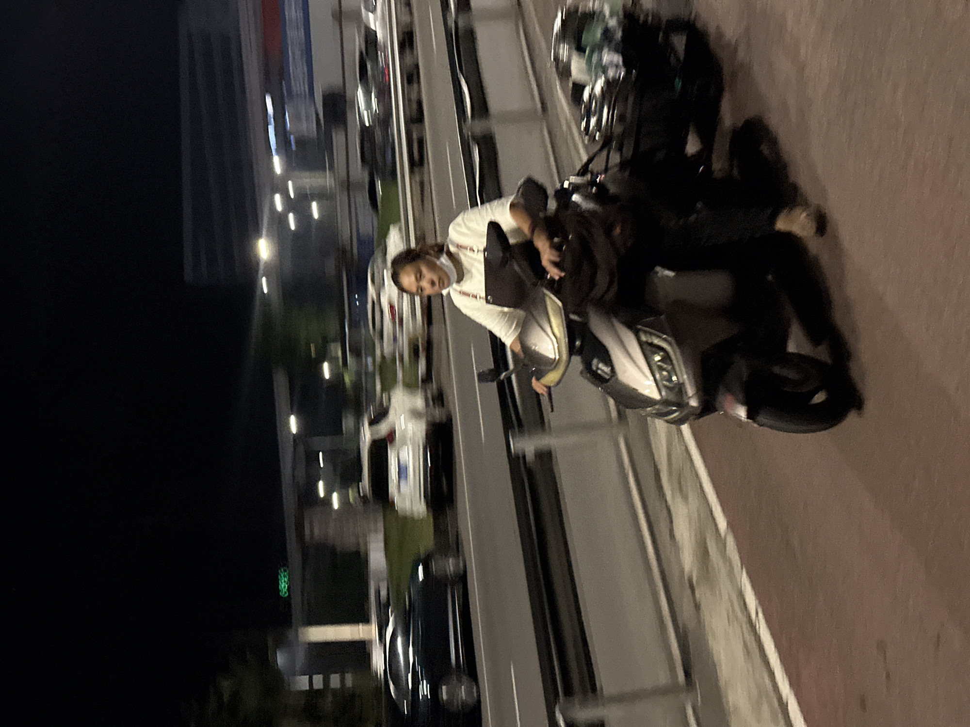 A medium shot captures a woman riding a scooter at night in the Chongqing High-tech Zone, China. The scooter is moving from left to right, and the image has motion blur. The woman is wearing a white t-shirt and a dark apron over it, with a white mask around her neck. She has dark hair pulled back. The scooter is a light color with a dark seat. Behind her, a metal rack on the back of the scooter is loaded with items covered in a blue and green cloth. The scooter is on a reddish-brown paved surface, possibly a bike lane, with a white line marking its edge. To the left of the paved surface is a gray road with cars in motion, indicated by the blur. A dark-colored car and a white SUV are visible on the road. In the background, there are streetlights and the blurry outlines of buildings. A sign with green lights is visible in the upper left corner. The overall scene suggests urban nighttime transit in a commercial or industrial area.