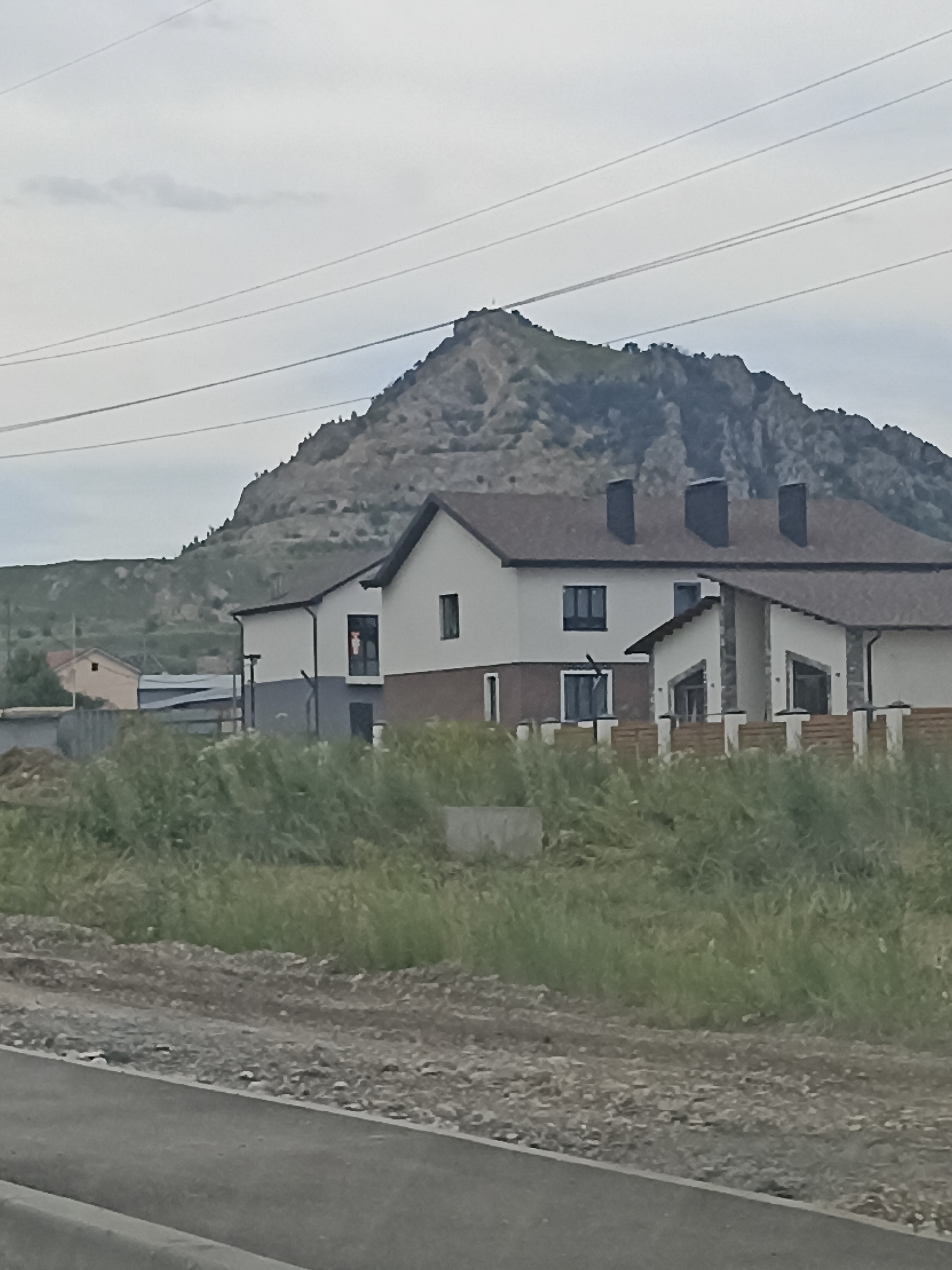 In Vinsady, Russia, a two-story residential building with a gray and white facade and a brown roof is pictured in the foreground, partially obscured by tall, green grass. The house has dark-framed windows. Behind it, a single-story building with a beige exterior is visible. In the background, a rugged, barren mountain rises under an overcast sky. Several power lines stretch across the upper portion of the frame, above the mountain. The foreground also shows a gravelly road and a paved surface.