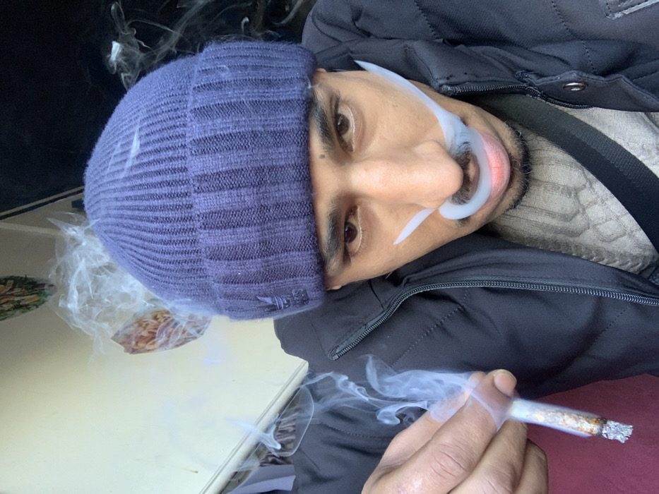 A man with a blue beanie and dark jacket faces the camera, holding a lit cigarette that emits smoke. He wears a gray patterned shirt under the jacket. Additional smoke surrounds his head and face. The image is slightly tilted.