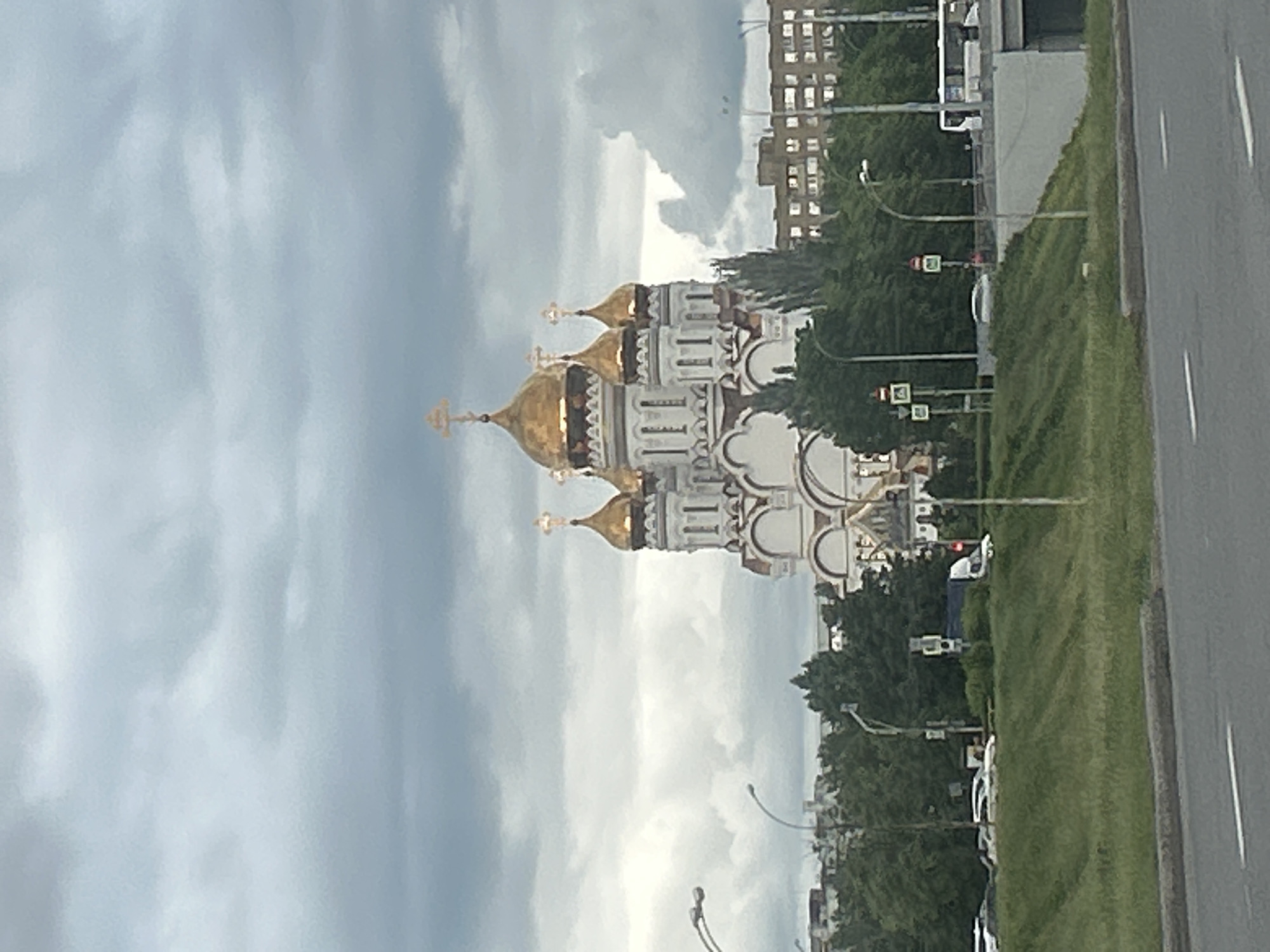 A low-angle, tilted outdoor shot depicts a white and gold Orthodox church with onion domes, juxtaposed against a cloudy sky, trees, and a grassy median. In the background is a building and a road to the right. The location is likely in Приморский, Russia, given the architectural style of the church.