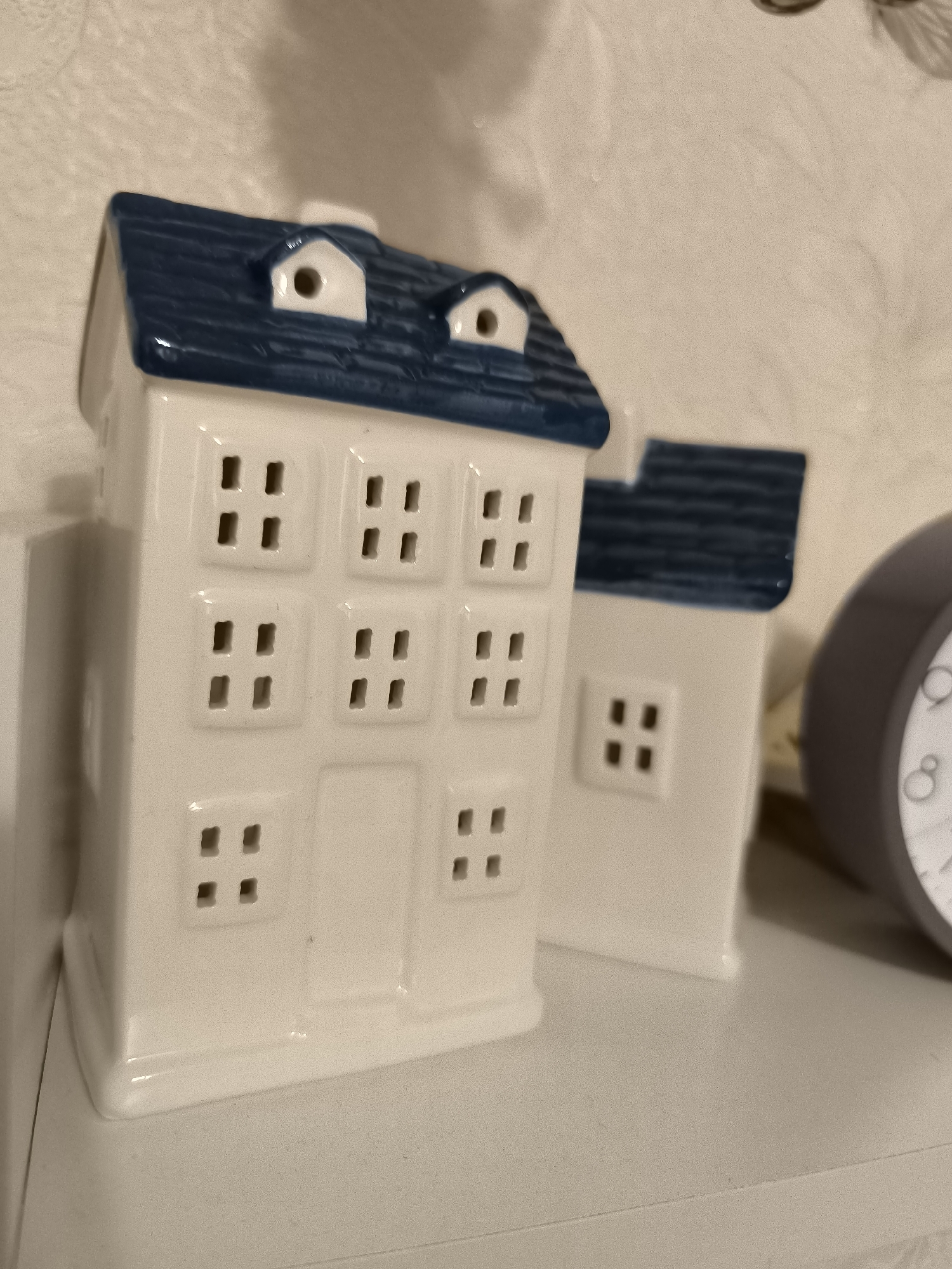 The image shows two decorative ceramic houses with blue roofs and white walls. The houses are placed on a white shelf. The house in the foreground is taller and has multiple square-shaped windows with four smaller squares inside each. The house in the background is partially visible and appears to be similar in design. To the right of the houses, a portion of a gray clock is visible. The background wall is textured and light-colored. The scene is indoors, and no people are present. The location in the city of Horishni Plavni, Ukraine, is not discernible from the image.