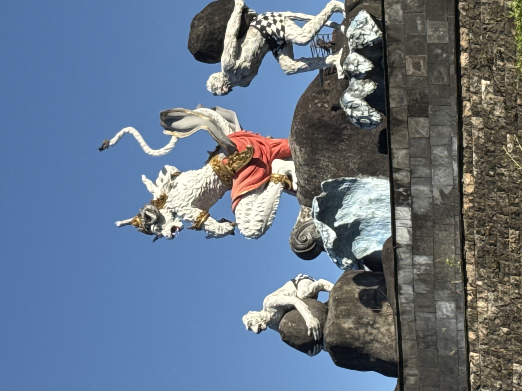 The image captures a portion of a stone statue, likely located in Denpasar, Indonesia, given the style of sculpture and architecture. The sculpture depicts a white, fantastical animal, possibly a lion or horse with ornate headgear and a billowing tail, ridden by a figure in a red robe. Above the animal, a figure with a checkered pattern on its torso appears to be lifting a large, dark stone. Below, another similar figure clings to a dark, rocky surface. The statues are positioned against a clear blue sky and are part of a larger structure with rough, textured surfaces and a central portion constructed from rectangular stone blocks.