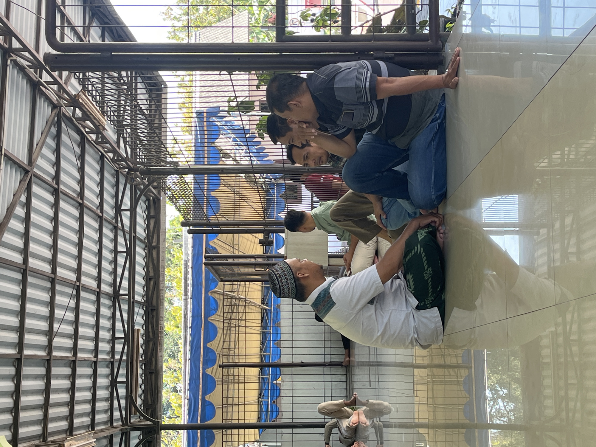 In Singosari, Indonesia, a group of men are gathered on a tiled floor under a corrugated metal roof supported by metal beams and pillars. The scene appears to be an open-air shelter. One man in the foreground wears a white shirt and a green patterned sarong, with a kufi on his head. He is seated, facing left, with his hands resting on his lap. To his right, three other men are seated in a group. The man on the far right wears a black and grey striped shirt and blue jeans. He is leaning back slightly, with his arm resting on his knee and his hand on the floor. The man next to him, in the center of the group, wears a light green shirt and has his hand to his face as if listening or contemplating. The fourth man in the group, on the far left, wears a light green shirt. In the background, two other individuals are visible, one lying down with his legs extended, and another seated. Beyond the shelter, a blue and yellow awning is visible, and trees and a building can be seen in the distance. The floor is highly reflective, mirroring the roof and the surrounding environment.