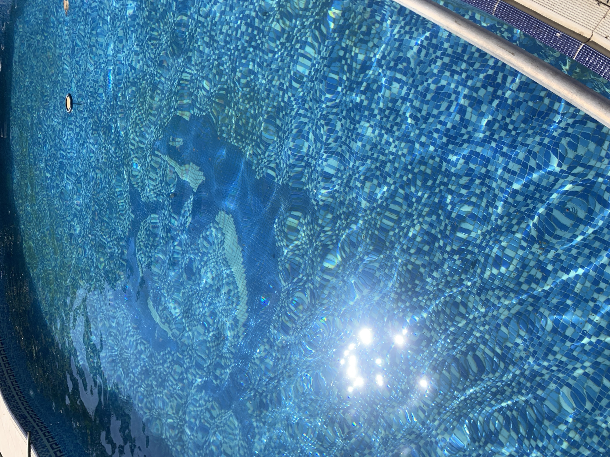 An overhead, medium shot captures the sunlit surface of a blue tiled swimming pool. The water is clear and rippled, with sunlight glinting off its surface, creating bright, reflective spots. The blue tiles beneath the water form a checkered pattern of varying shades of blue. A silver-colored handrail runs diagonally across the lower right portion of the frame, with a textured white coping and blue tiled border visible along the edge of the pool. In the upper right quadrant of the pool, a small, dark object with a yellow ring floats on the surface. The scene is outdoors, likely during the day, given the strong sunlight. The pool appears to be an in-ground or above-ground structure with a decorative tile pattern along its inner wall, visible in the upper left corner of the image. The overall impression is of a clean, inviting swimming pool on a sunny day. The location in Sabunchu Raion, Azerbaijan, suggests a residential or recreational setting.