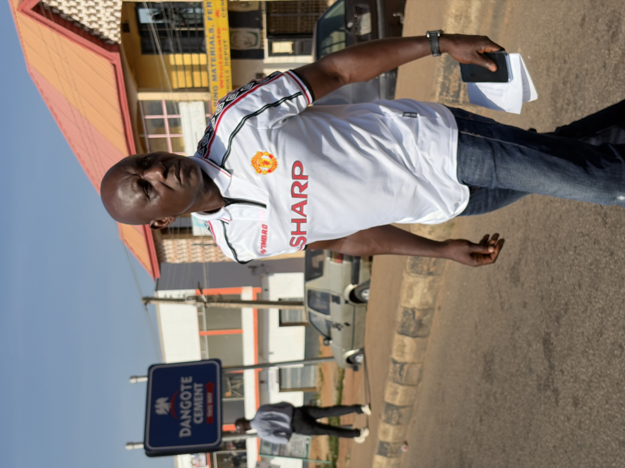 In Gura Top, Nigeria, a bald Black man walks along a paved road on a sunny day. He is wearing a white Manchester United soccer jersey with red and black trim and blue denim jeans. He is carrying a black smartphone and a stack of white papers in his right hand. A blue and white street sign for "DANGOTE CEMENT" is visible behind him. A light-colored car is parked on the side of the road in the background, and a building with an orange roof is also visible. Another Black man in a light blue shirt and dark pants is walking away from the camera in the background.