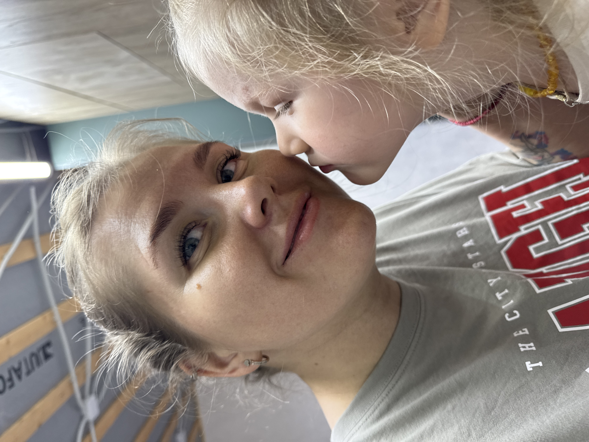 A young child with blonde hair is kissing the cheek of a woman with blonde hair. The woman is wearing a gray t-shirt with red and white lettering that reads "THE CITY STAR". The background shows a ceiling and wall, suggesting an indoor location. The city mentioned is Ферма, Russia. The photo appears to be taken at close range, focusing on the interaction between the two individuals.