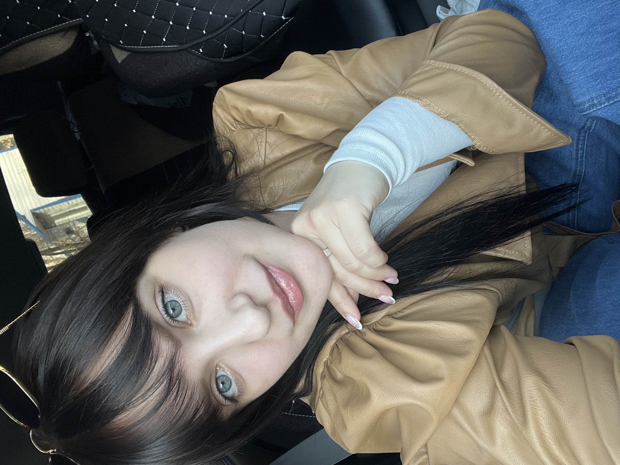 A young woman with long, dark hair and striking light blue eyes is featured in a close-up portrait, appearing to be inside a car. She wears a light brown, possibly faux leather, jacket over a white long-sleeved top, and blue jeans. Her chin is gently rested on her right hand, which displays a ring with a square-cut stone on her ring finger and manicured nails with a French tip. A pair of gold-framed glasses is partially visible near her left temple.
The background elements include a black quilted car seat cover on the upper left and dark interior components of a vehicle. Through the window on the far left, bright daylight illuminates what appear to be industrial structures or buildings, suggesting it is daytime. The woman has a calm and pleasant expression with a soft smile, looking directly at the camera. The setting is a vehicle in Chaikovsky, Russia. There is no visible text in the image.