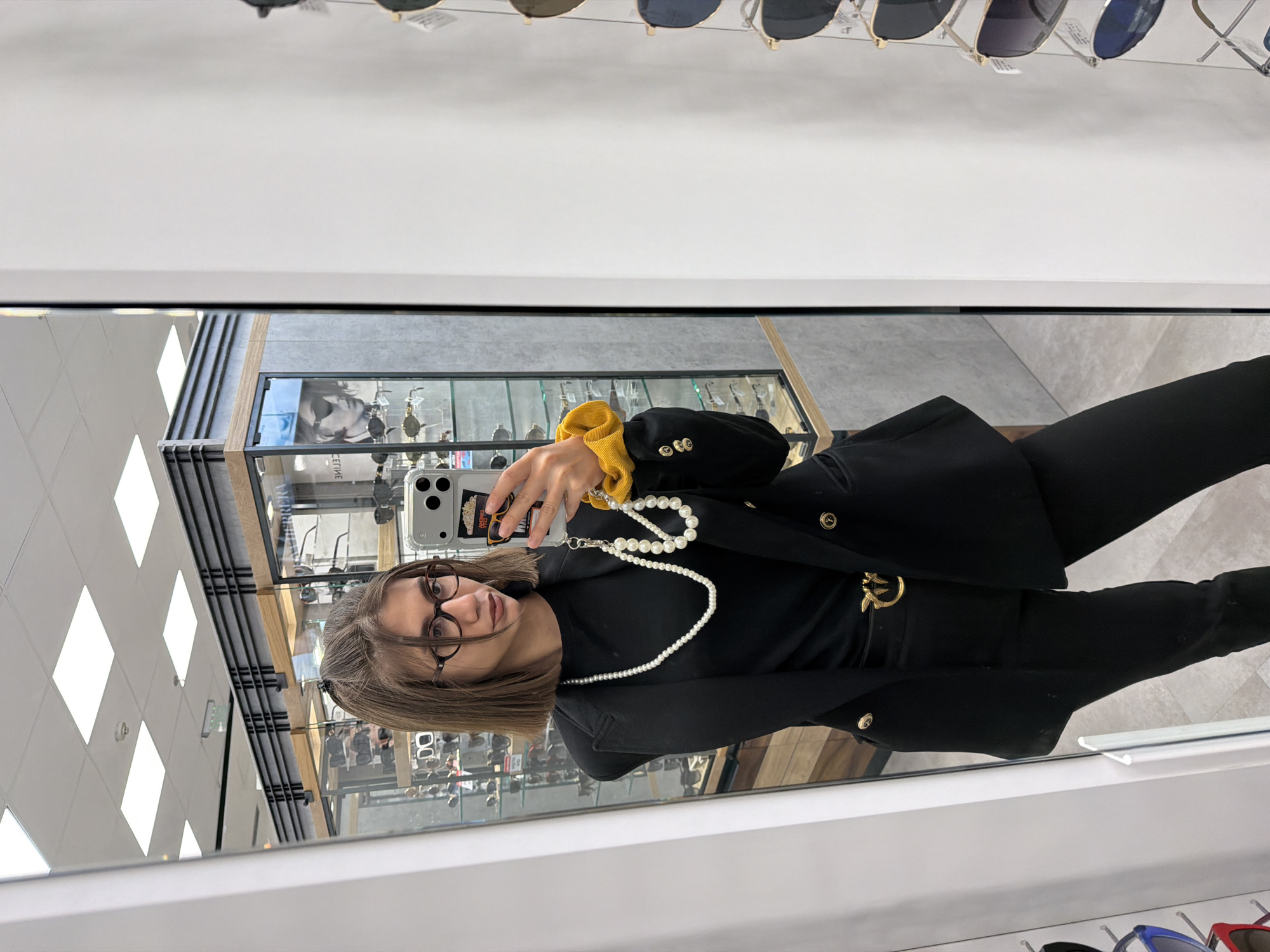 A young woman stands in front of a mirror, taking a selfie. She is wearing a black blazer, black pants, a black top, glasses, and a pearl necklace. Her hair is pulled back in a ponytail. She is holding a smartphone with a popcorn-themed phone case. In the background, there are displays of glasses and sunglasses. The location appears to be an optical shop.