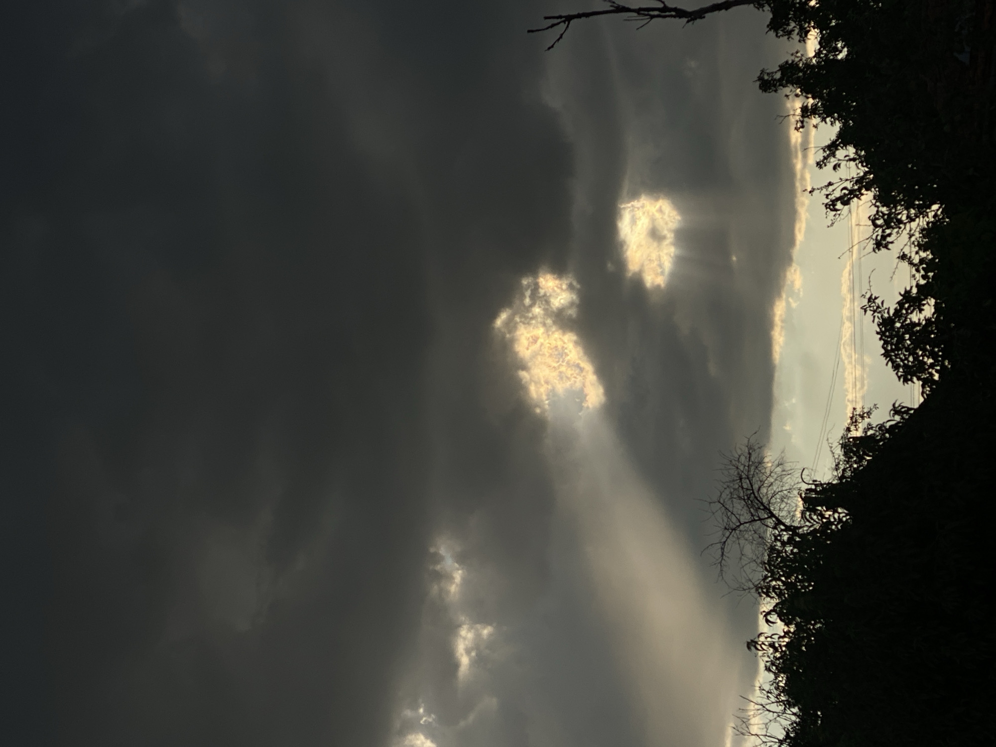 The image depicts a dramatic sky with dark, ominous clouds. Shafts of sunlight, known as crepuscular rays, pierce through breaks in the clouds, illuminating patches of the sky with a golden glow. In the foreground, the silhouettes of trees and branches are visible against the lighter sky, obscuring the horizon. The overall impression is one of a stormy or impending storm sky with a touch of dramatic lighting. The location is identified as Cairaclia, Moldova. No people or specific actions are observable in the image.
