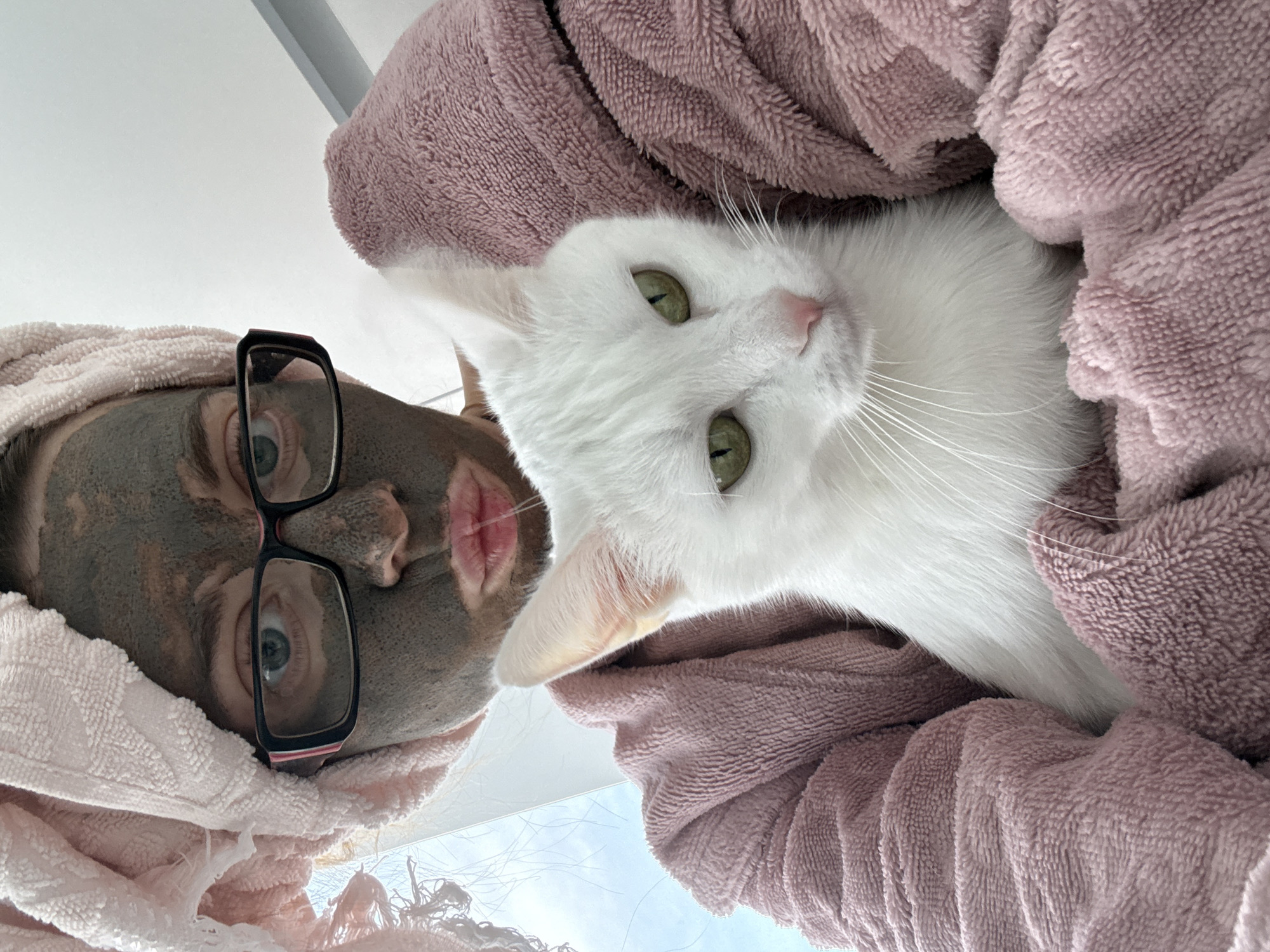 In this selfie, a person with a pink towel wrapped around their head and a clay face mask on their face is holding a white cat. The person is wearing glasses and a pink bathrobe. The cat has green eyes and is looking at the camera. The background is out of focus and appears to be indoors.