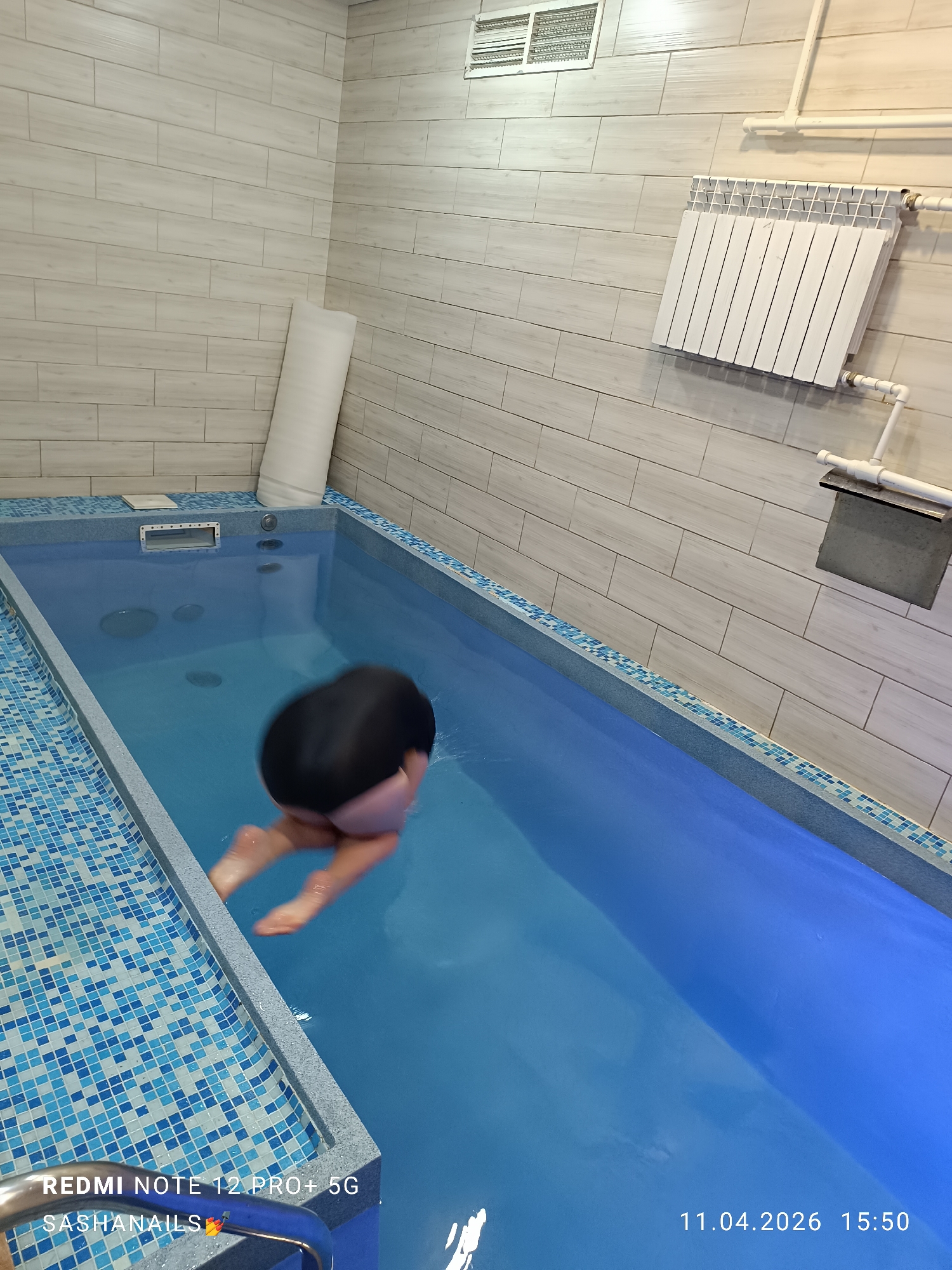 The image depicts an indoor swimming pool, with a person partially submerged and blurred as if in motion. The pool is rectangular, lined with blue mosaic tiles. The water is light blue and reflects the light.
The walls of the room are tiled with light gray horizontal rectangular tiles. There's a white, cylindrical object standing against the wall behind the pool. Above the pool, there are pipes and a white radiator mounted on the wall. To the right of the radiator is a grey rectangular object attached to the wall, possibly part of the plumbing.
The person in the pool appears to be diving or entering the water headfirst, with their head and back visible above the water.
The image includes a date stamp in the bottom right corner, "11.04.2026 15:50," and text in the bottom left corner, "REDMI NOTE 12 PRO+ 5G SASHA NAILS".
The location is Chaikovsky, Russia.