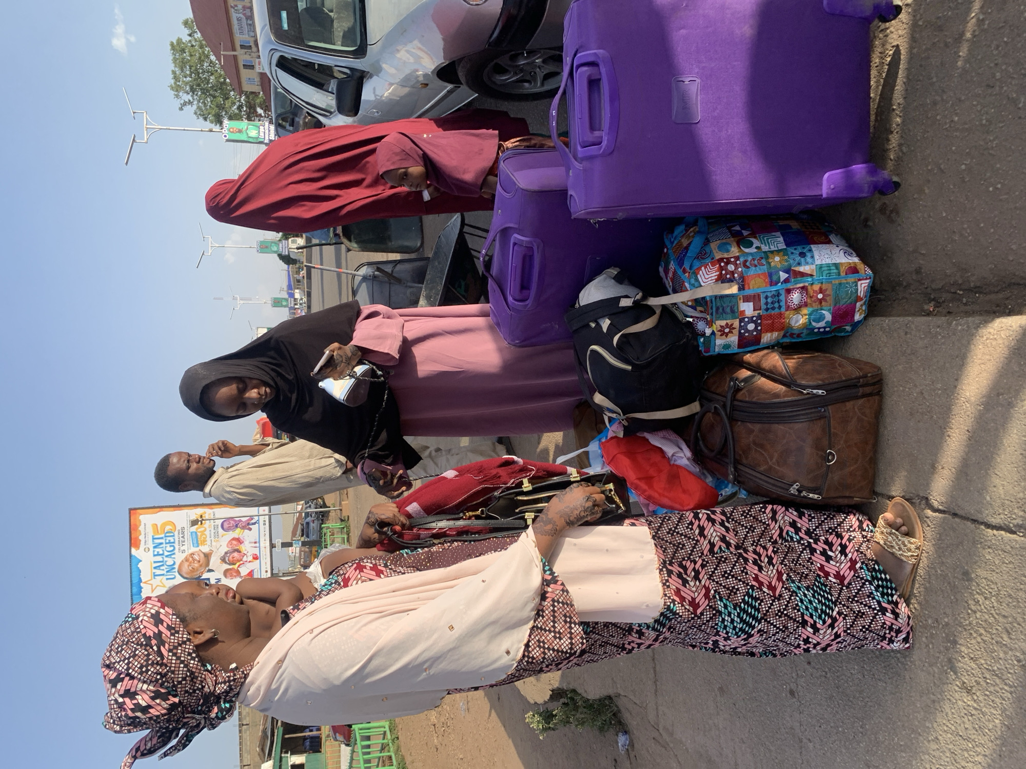 The image depicts a group of people and luggage on a street in Gura Top, Nigeria. An older woman in a patterned headwrap and dress sits near a collection of luggage: two purple rolling suitcases, a colorful patterned bag, and a brown duffel bag. Another woman in a black hijab and pink dress stands nearby, looking at a cell phone. A third woman in a red hijab stands behind the luggage. A man in a light-colored shirt is visible in the background, smoking a cigarette. The backdrop includes a silver car, streetlights, buildings, and a billboard advertising "Talent Uncaged." The overall impression is of a roadside scene with travelers and their belongings.