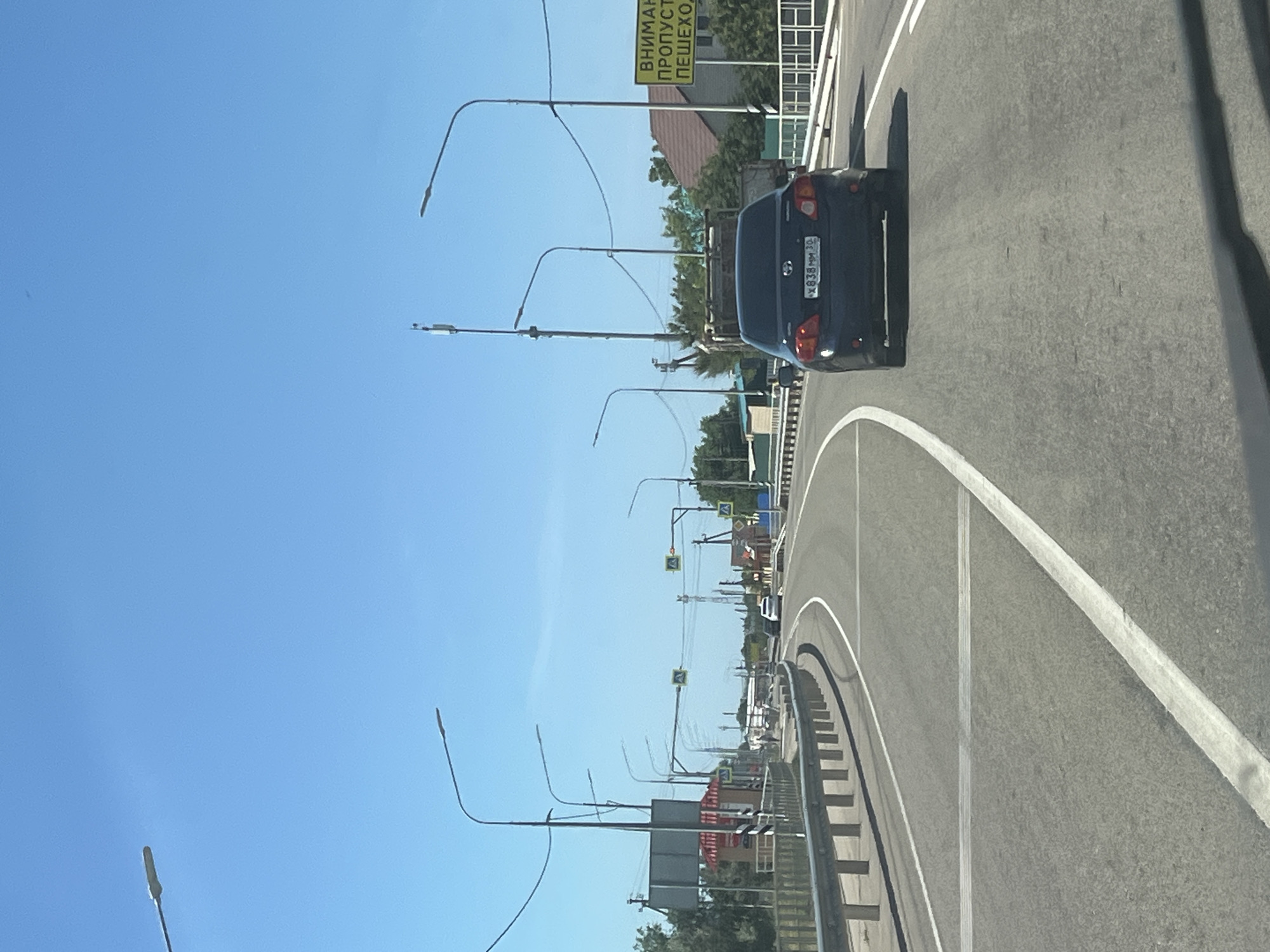 The image depicts a view from a vehicle traveling on a paved road with a clear blue sky overhead. The road curves to the left, bordered by a metal guardrail on the left and a white line painted on the asphalt. Streetlights are visible along the road. In the distance, there are buildings, trees, and more streetlights. A dark blue sedan is in front of the camera vehicle. A yellow sign with Russian text is visible on the right side of the road. The location is identified as Bakhtemir, Russia.