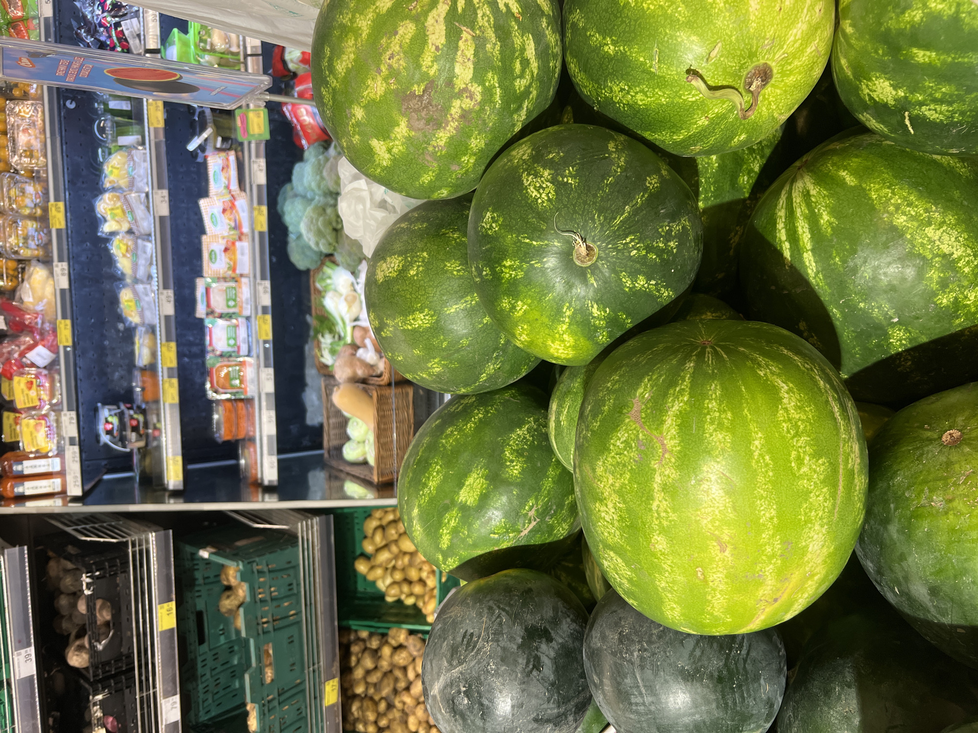 A close-up, eye-level shot shows a display of watermelons in a grocery store in Odesa, Ukraine. The watermelons are predominantly green with lighter green striping, and some darker, almost black watermelons are also visible. They are piled in a large display, with some partially obscured by others. The lighting suggests an indoor environment, likely a supermarket with artificial lighting. The background is slightly out of focus but shows shelving with various food items, including produce like potatoes and broccoli, and packaged goods. The overall impression is of a fresh produce section.