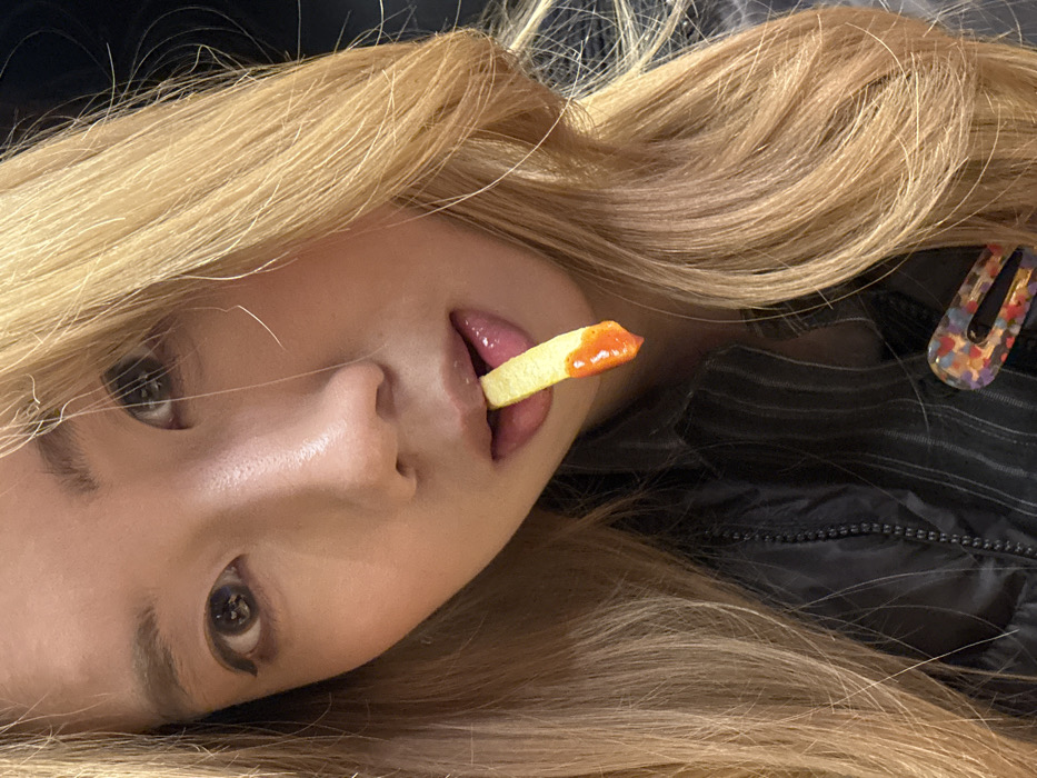 A blonde woman lies on her side, with a french fry covered in red sauce between her lips. Her eyes look upward. Her hair is styled loosely around her face and body. A colorful hairpin decorates the side of her head. She is wearing a dark jacket or shirt, partially obscured by her hair. The image is rotated slightly, giving a tilted perspective.