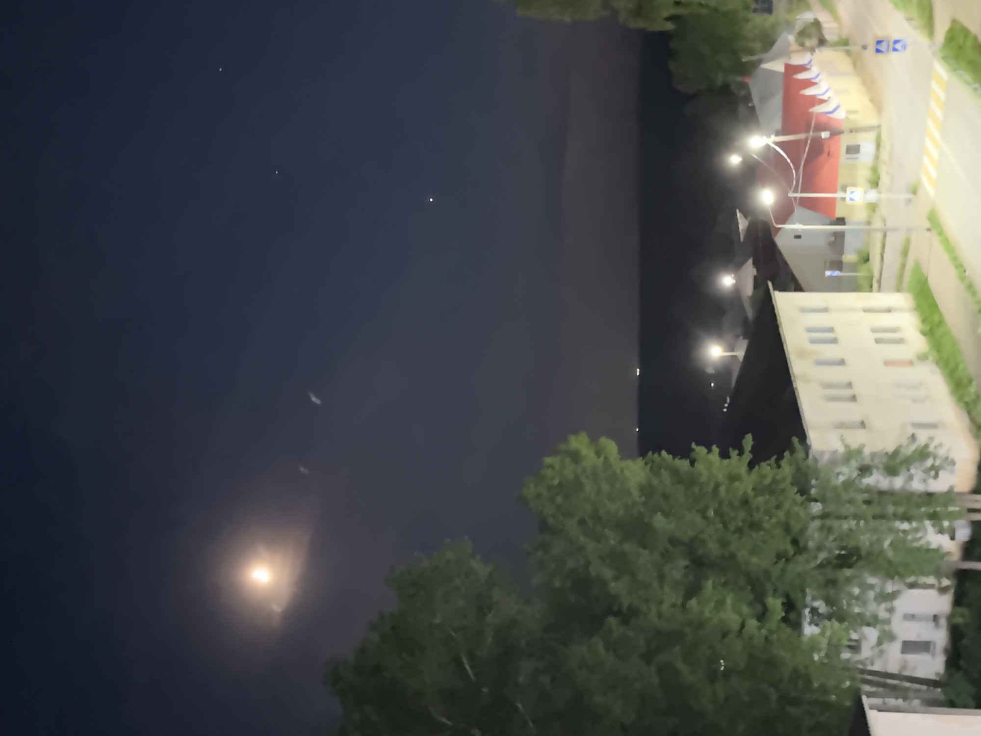 The image shows a night scene in what appears to be a residential area. The sky is dark with the moon visible. There are buildings lit by street lights, trees, and a road. The road has white and yellow lines indicating lanes and crosswalks. There are also Russian flags. Based on the provided information and the presence of the Russian flags, this is a street in Chaikovsky, Russia.