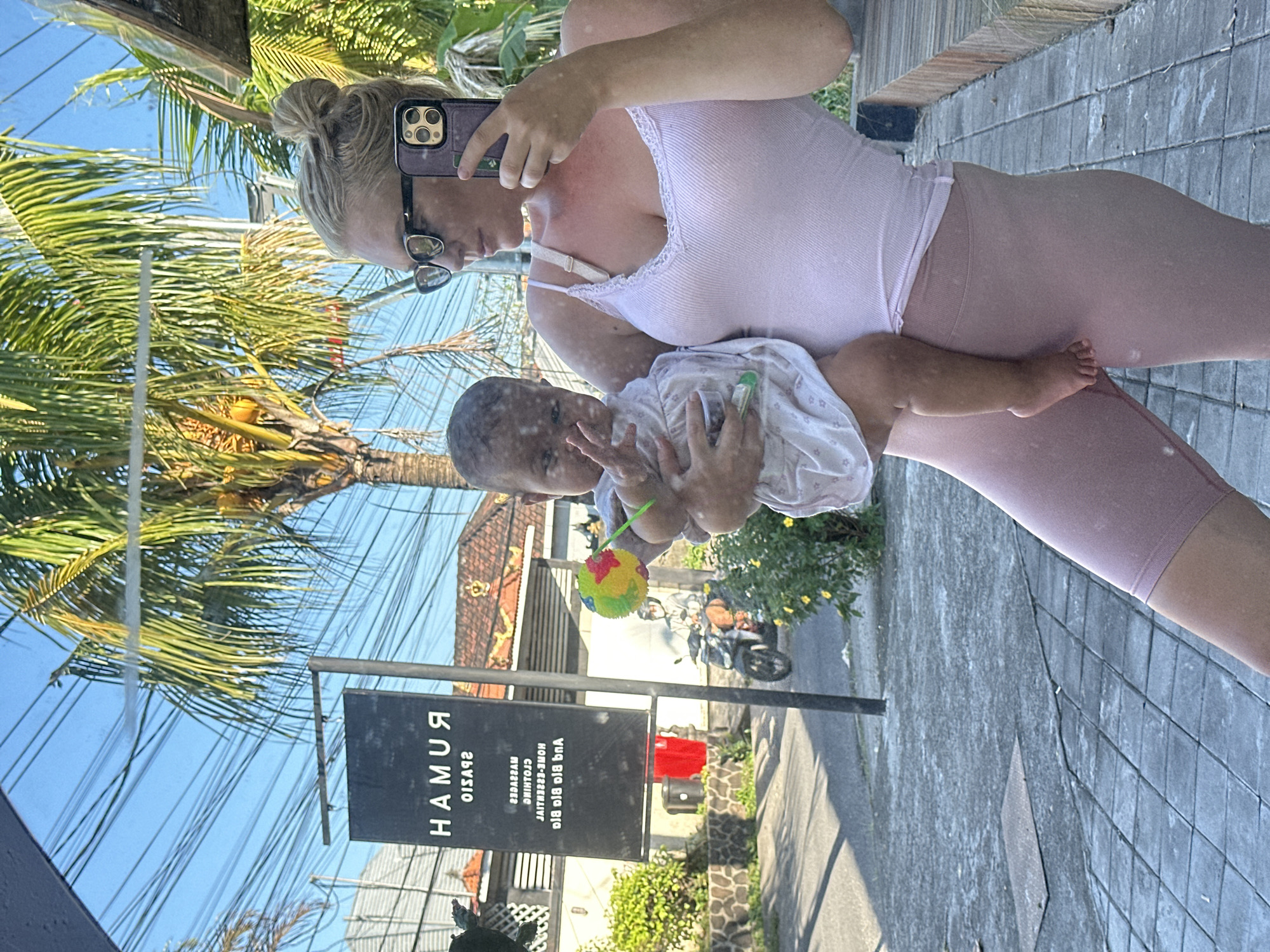 A reflective outdoor scene shows a woman holding a baby, possibly a self-portrait. The woman has her hair in a bun and is wearing sunglasses, a light pink tank top, and matching biker shorts. She is holding a phone in her left hand, presumably to take the picture. The baby is wearing a light-colored outfit and is holding a colorful toy.
The reflection shows a tropical outdoor setting. In the background, there's a sign that reads "RUMAH PHYSIO" in reverse, indicating a physiotherapy center. The surroundings include palm trees, buildings with tiled roofs, and a glimpse of a street with a parked motorbike.
Based on the sign and tropical setting, the location is likely in North Kuta, Indonesia. The reflection suggests the photo is taken at a glass surface or a reflective panel.